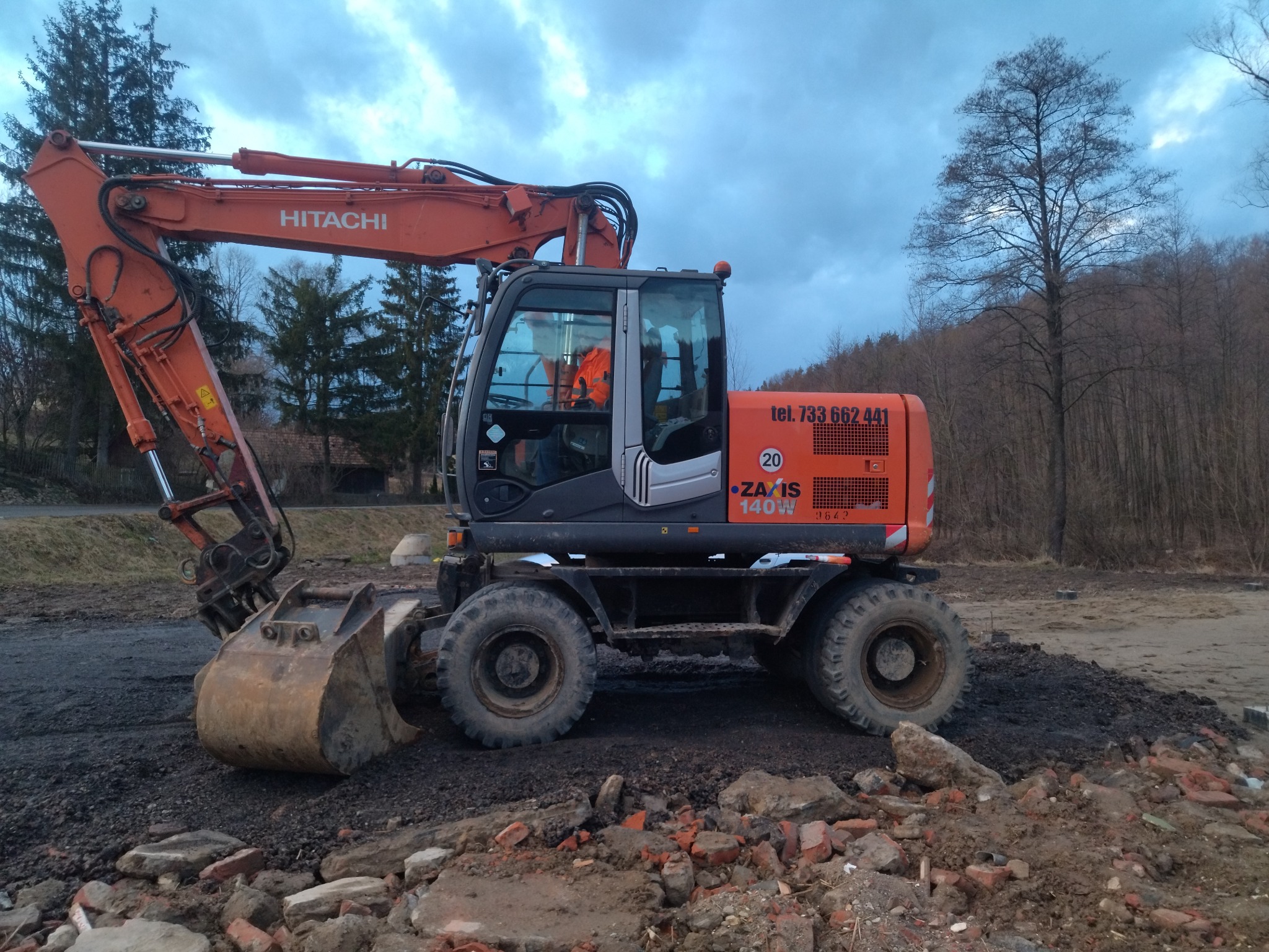 Pomarańczowo-szary kołowy koparka Hitachi Zaxis 140W na nierównym terenie z gruzem i ziemią, widoczny operator w pomarańczowej kamizelce, drzewa w tle.