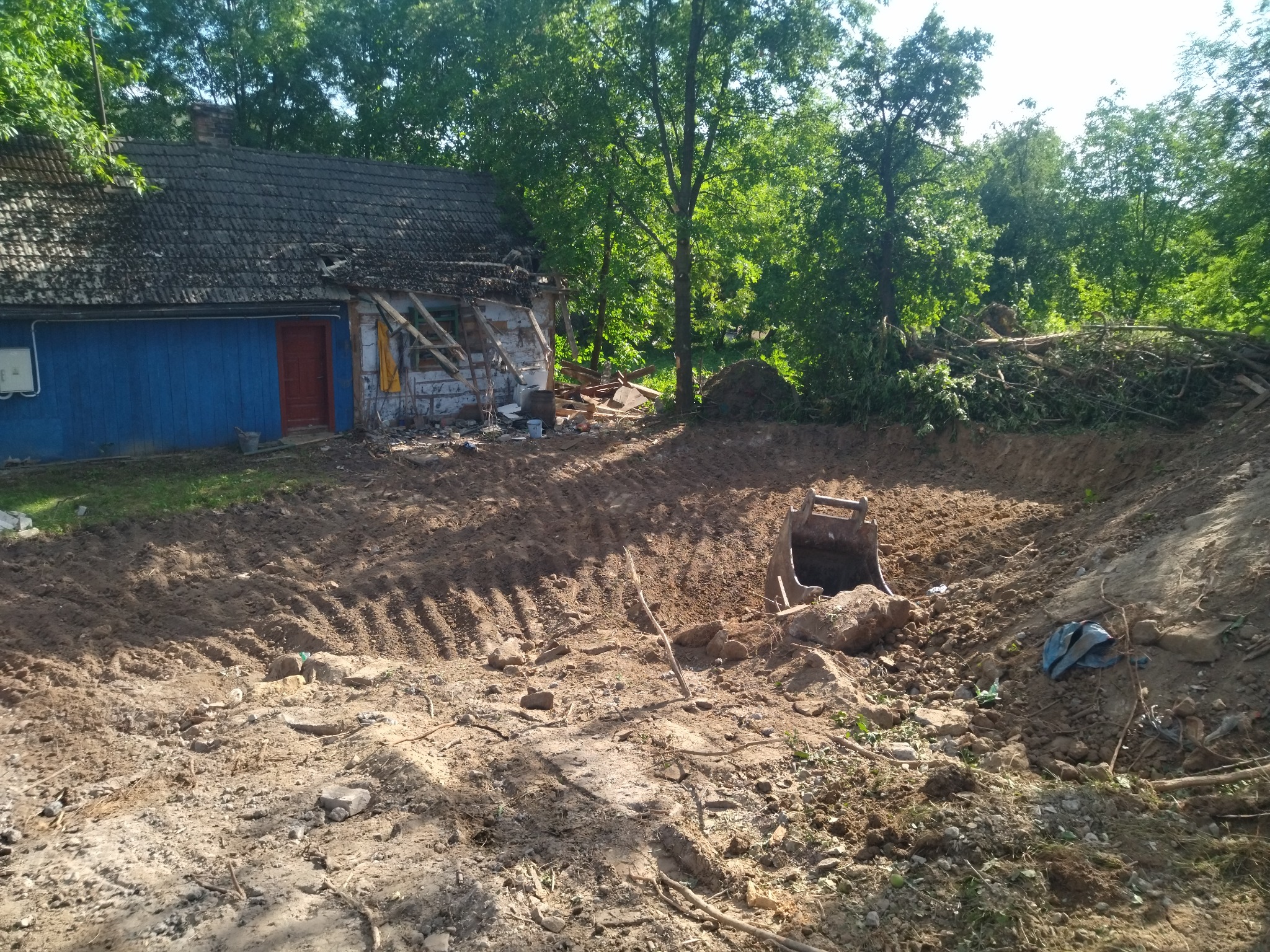 Szeroki wykop w ziemi z widoczną łyżką koparki, w tle częściowo zrujnowany budynek z niebieską ścianą i stertą gruzu, otoczony drzewami.