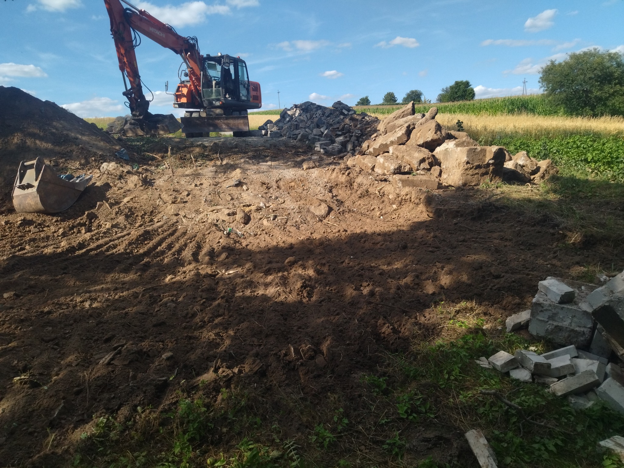 Pomarańczowa koparka Hitachi na placu budowy, z wykopaną ziemią, stertą kamieni i gruzu, oraz polem kukurydzy w tle pod błękitnym niebem.