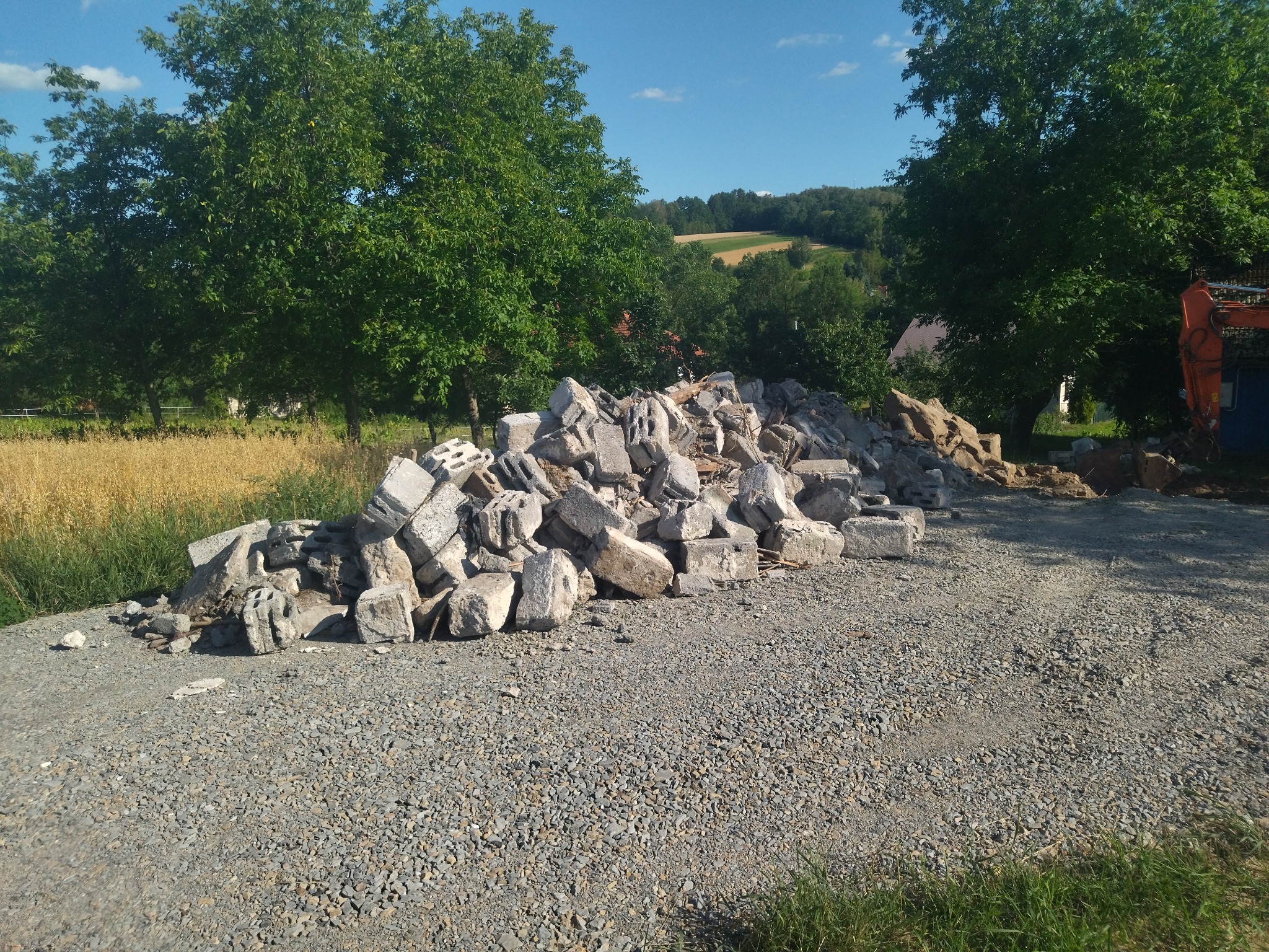 Kupa gruzu betonowego na żwirowej drodze, w tle pole zboża i drzewa, częściowo widoczny pomarańczowy sprzęt budowlany.