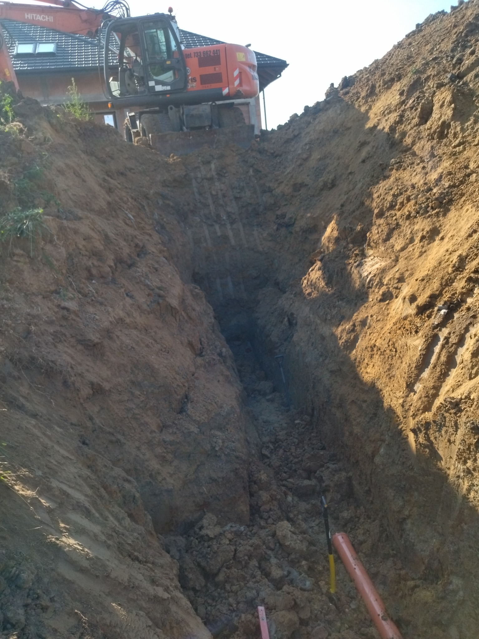 Pomarańczowa koparka Hitachi na skraju głębokiego wykopu w ziemi, z widocznym fragmentem budynku w tle i pomarańczową rurą na dnie wykopu.