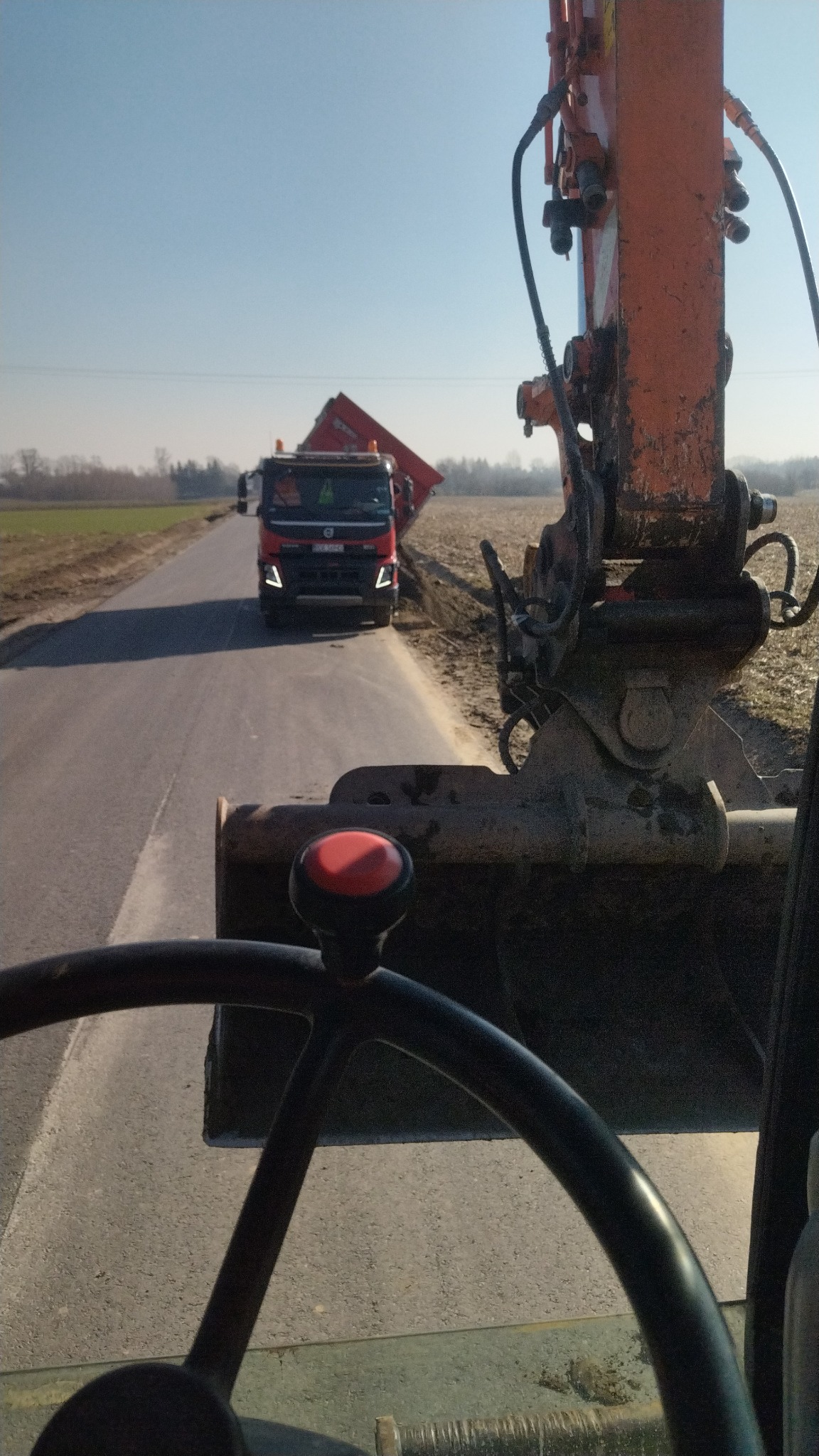 Widok z kabiny koparki na wywrotkę na drodze podczas prac ziemnych, ramię koparki po prawej stronie kadru, pole w tle.