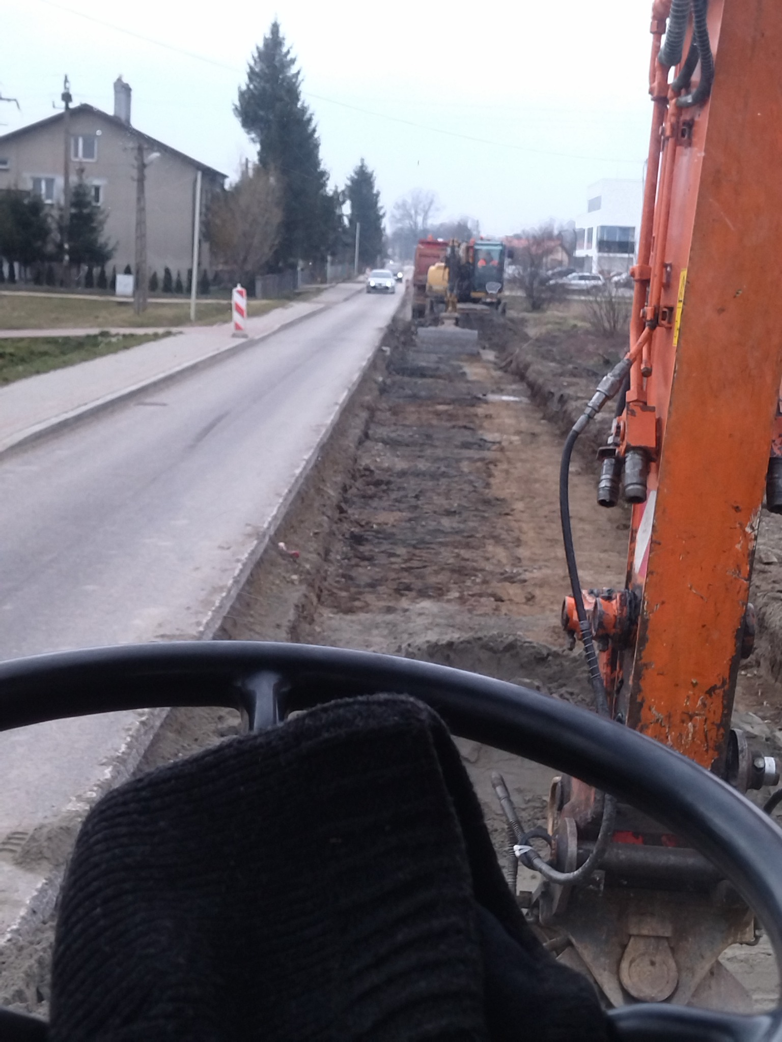 Widok z kabiny koparki na wykop wzdłuż ulicy, z widocznymi maszynami budowlanymi i ruchem samochodowym w tle, w pochmurny dzień.