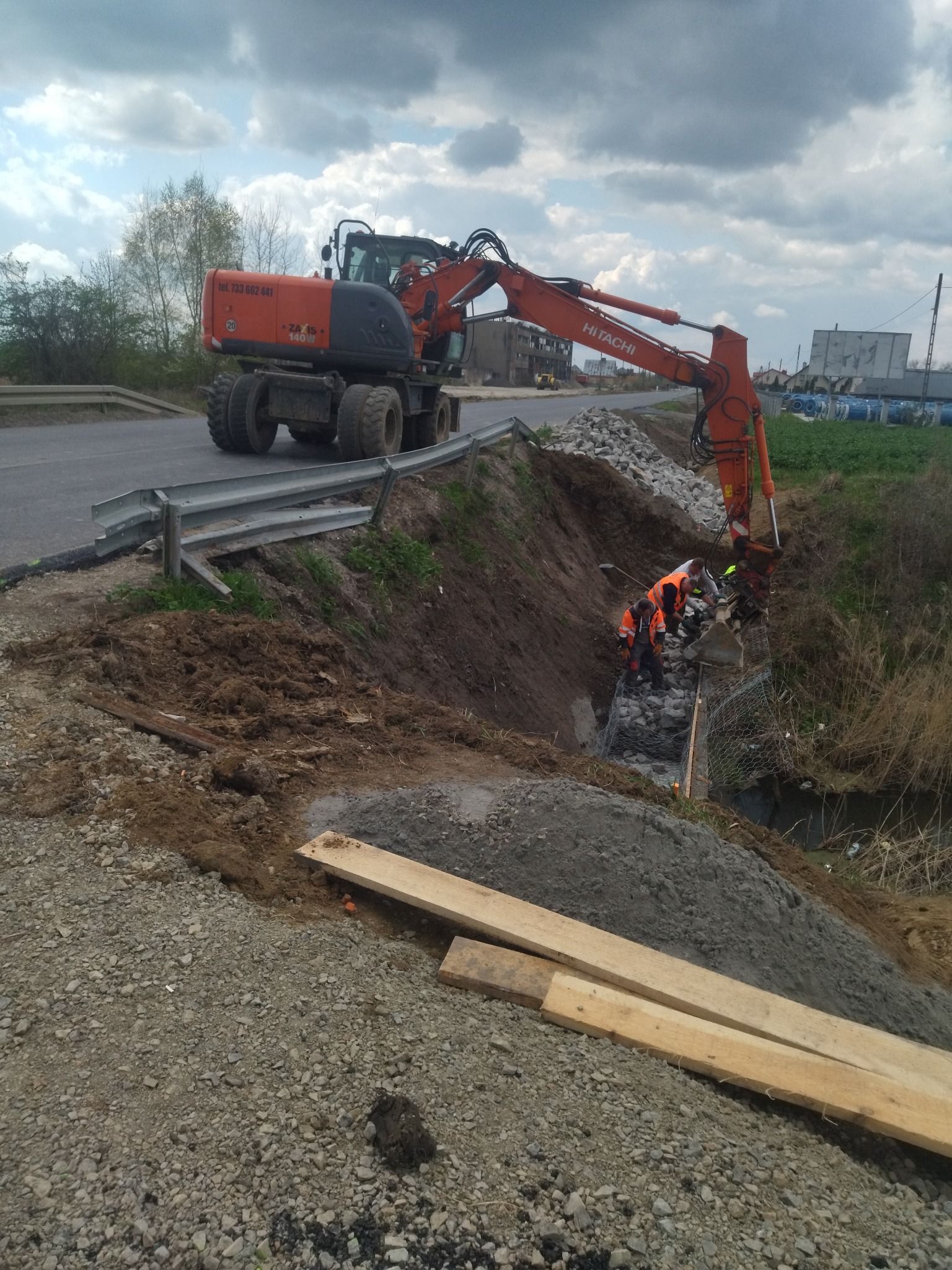 Pomarańczowa koparka Hitachi przy drodze, pracownicy w odblaskowych kamizelkach układają kamienie w siatce na zboczu, prawdopodobnie wzmacniając brzeg rowu.