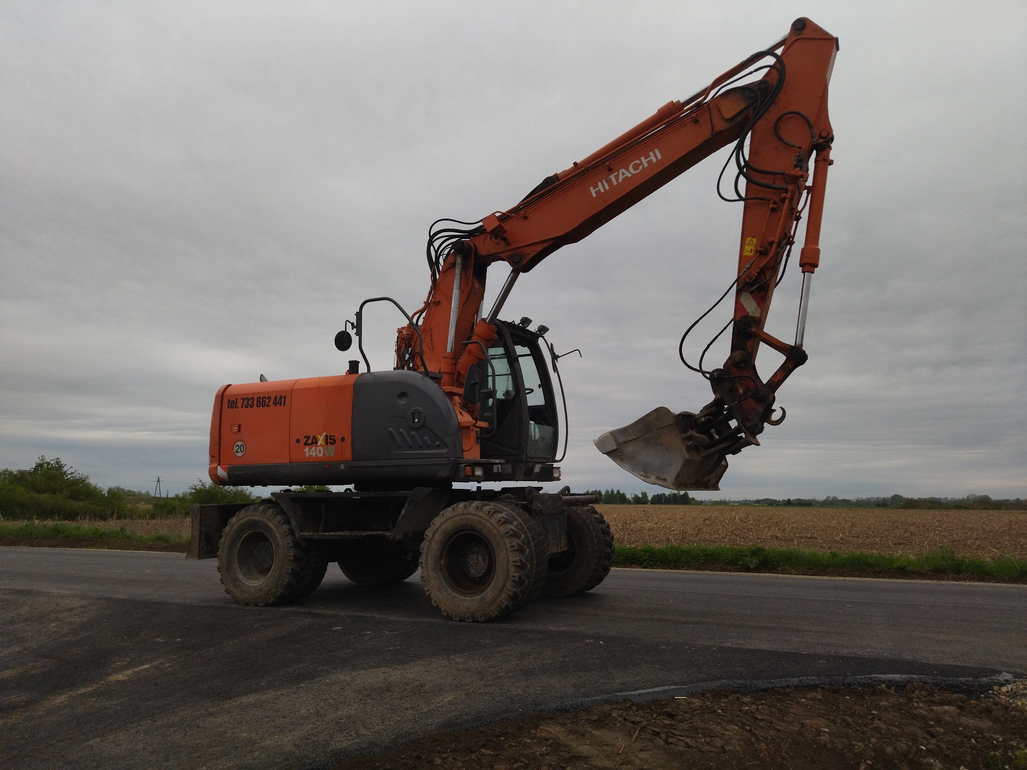 Pomarańczowo-szara koparka kołowa Hitachi Zaxis 140W stoi na asfalcie, ramię uniesione, łyżka lekko zabrudzona ziemią, w tle pole i pochmurne niebo.