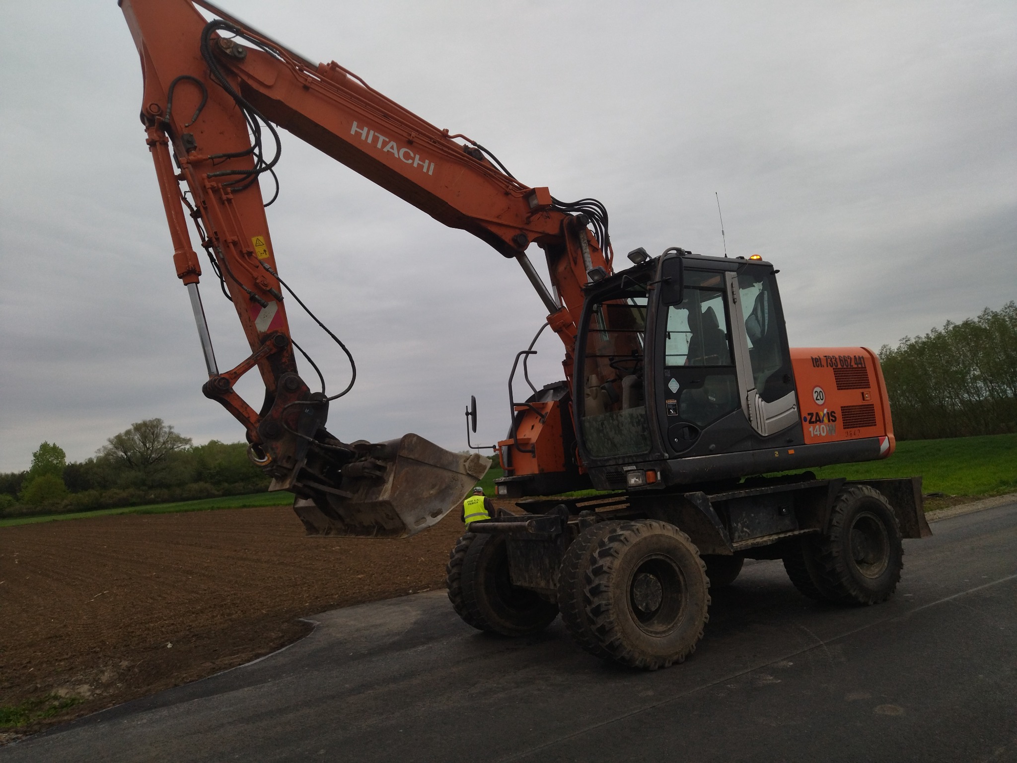Pomarańczowa koparka kołowa Hitachi Zaxis 140W na asfalcie, z widocznym polem uprawnym w tle i pracownikiem w żółtej kamizelce w pobliżu, pochmurne niebo.