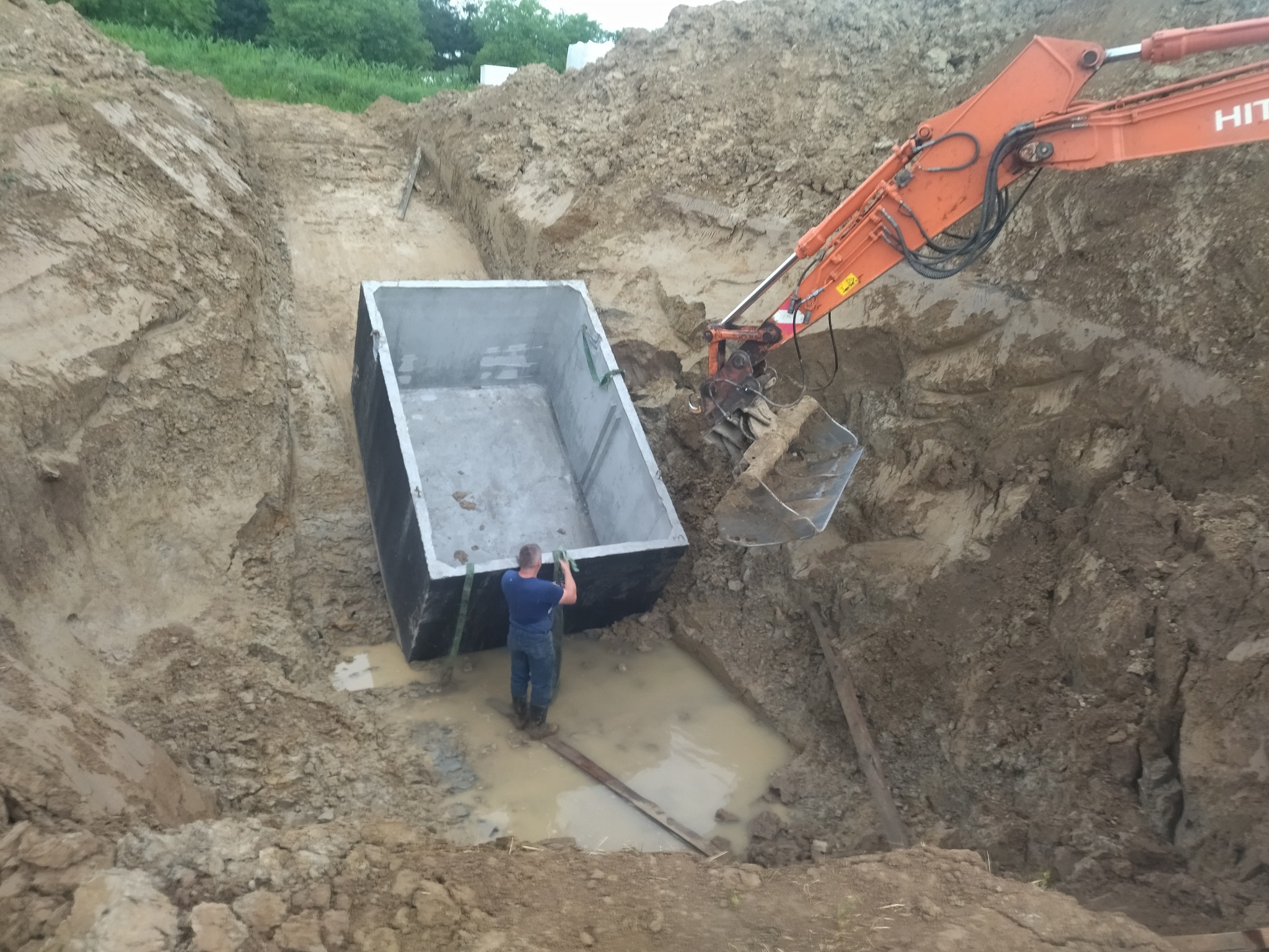 Pomarańczowa koparka HITACHI opuszcza betonowy zbiornik do wykopu wypełnionego wodą, pracownik w kaloszach pomaga w ustawieniu, ziemia wokół wykopu.