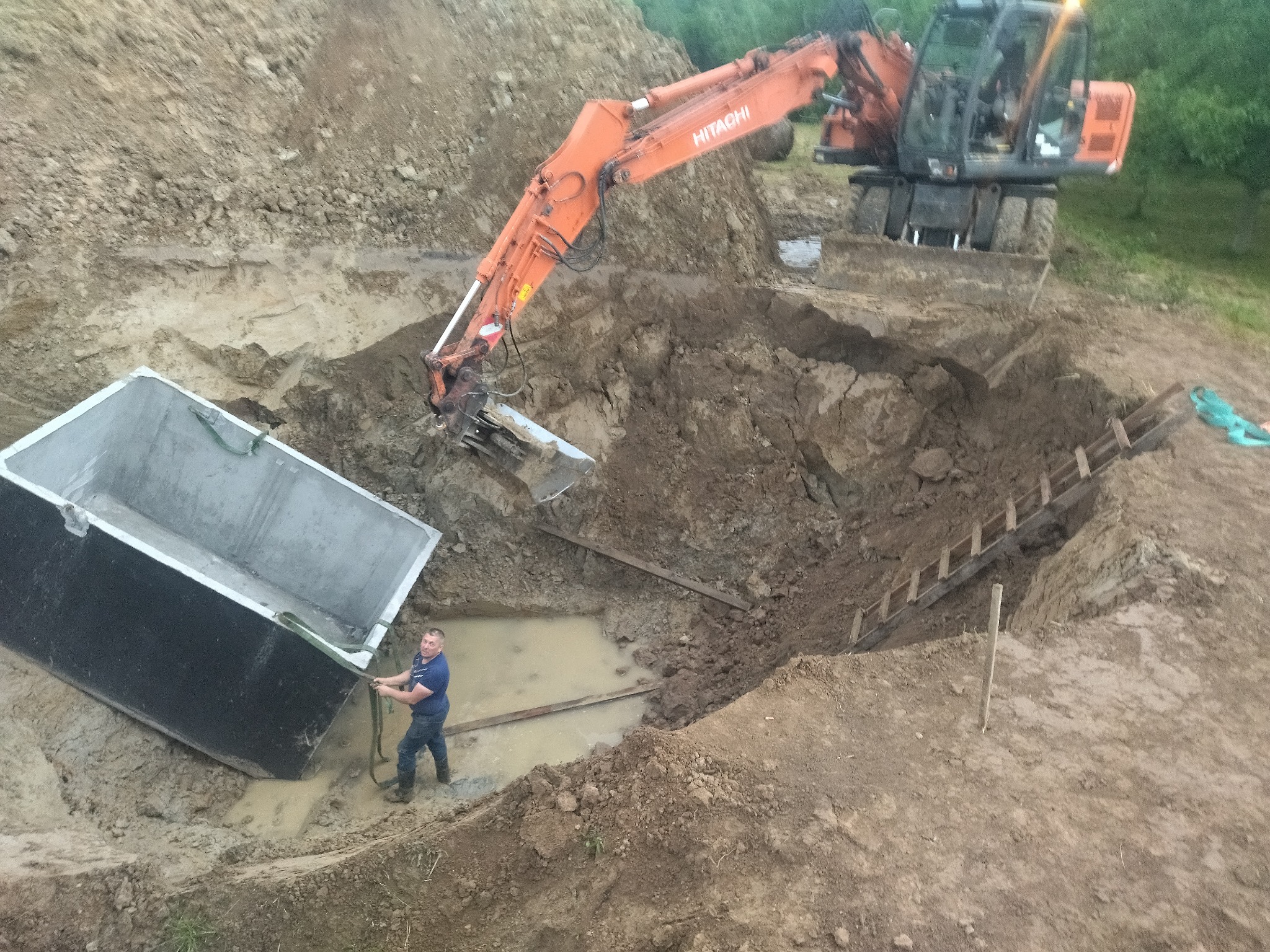 Montaż szamba w Pruchnik. Pracownik pomaga w ustawieniu betonowego zbiornika w wykopie, w tle pomarańczowa koparka Hitachi i strome zbocze wykopu zabezpieczone drewnianą drabiną.