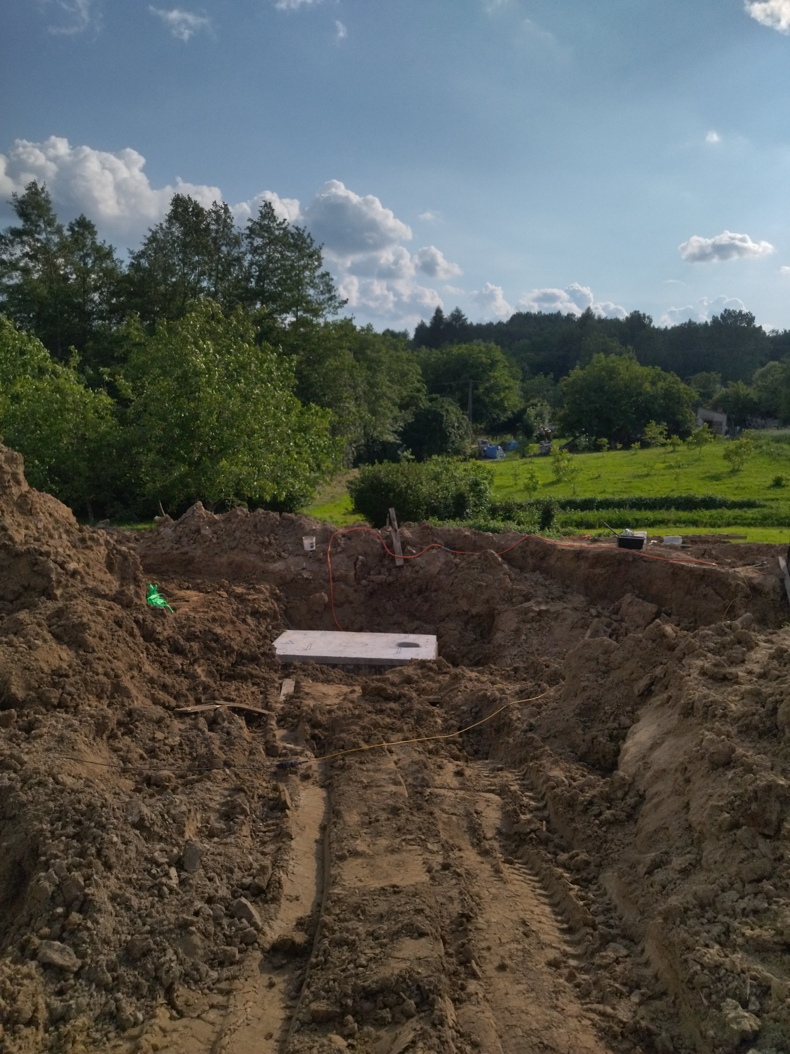 Wykop w ziemi z betonową płytą na dnie, widoczne ślady opon i przygotowane przewody, w tle zieleń drzew i łąk pod błękitnym niebem z chmurami.