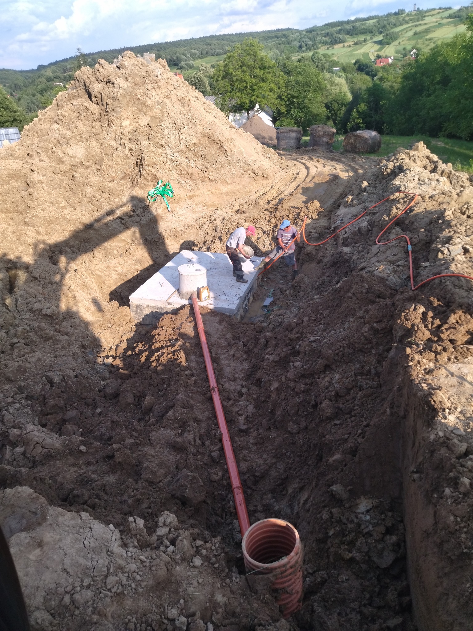 Montaż betonowego zbiornika, prawdopodobnie szamba, w wykopie. Widoczni dwaj pracownicy, rury i hałda ziemi. W tle zielony krajobraz wzgórz.