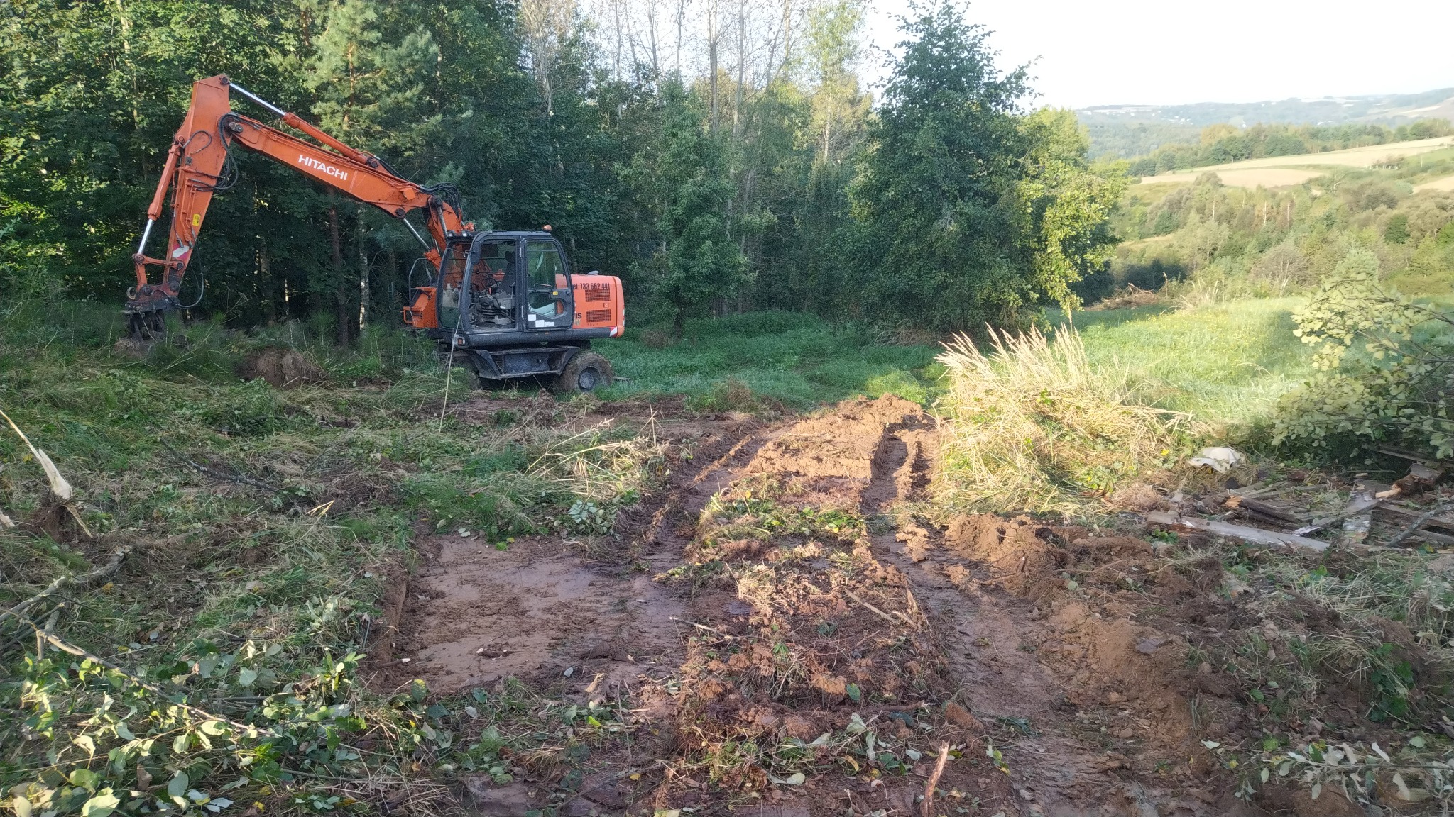 Pomarańczowa koparka Hitachi na nierównym, błotnistym terenie z wykarczowaną roślinnością, w tle zielony las i pagórkowaty krajobraz, widoczne ślady opon.