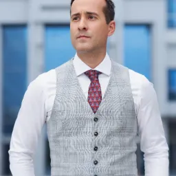 Mężczyzna w szarej kraciastej kamizelce, białej koszuli i wzorzystym krawacie, patrzy w dal na tle nowoczesnego budynku.