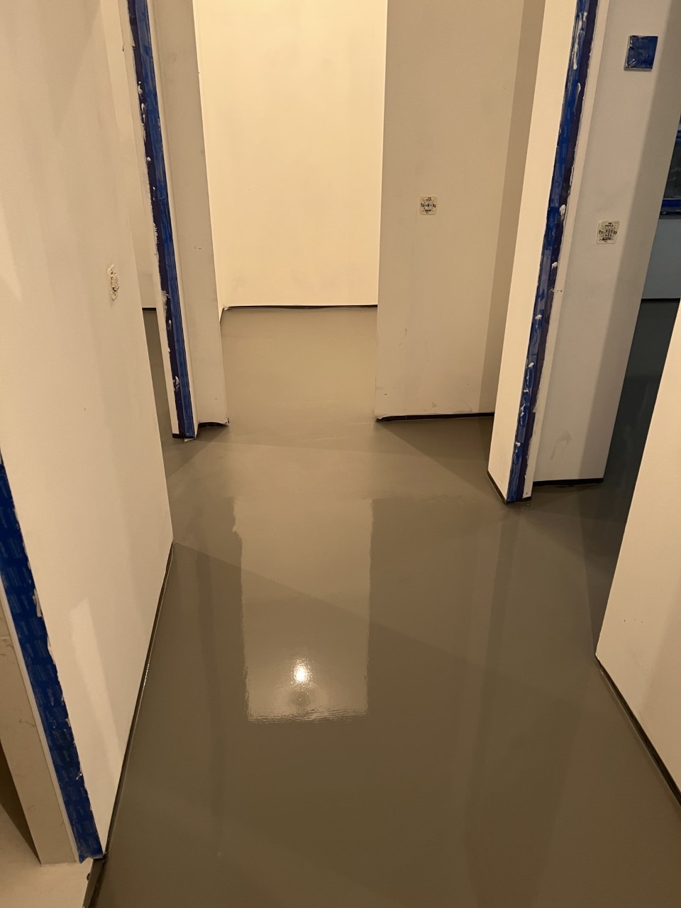 Świeżo wylana, gładka, szara posadzka mikrocementowa w korytarzu, zabezpieczona niebieską taśmą malarską przy ścianach i ościeżnicach.