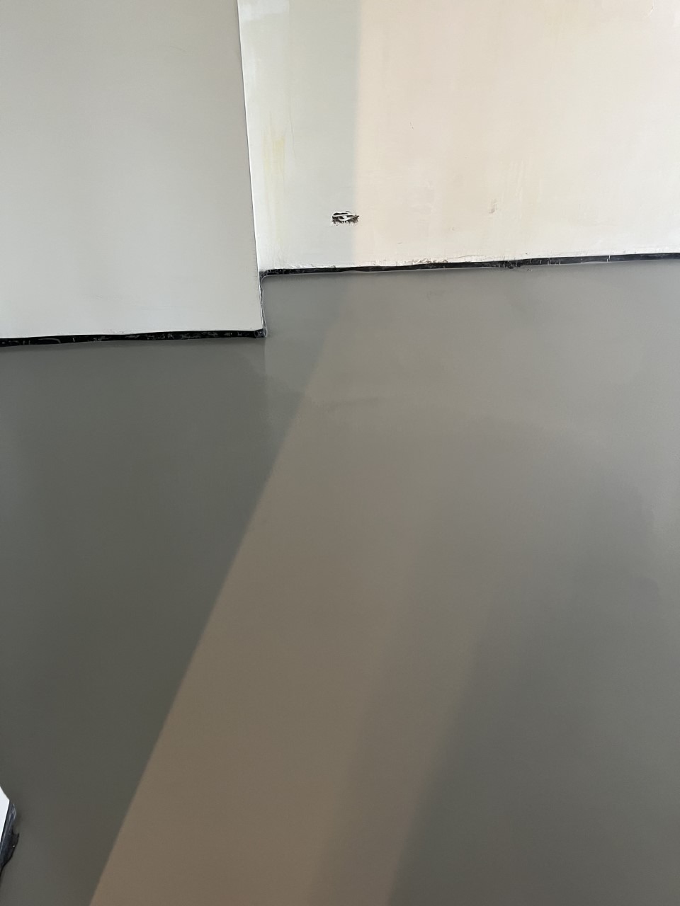 Świeżo wylana, gładka posadzka w szarym odcieniu, widoczne ściany przygotowane do malowania i czarne listwy przypodłogowe.