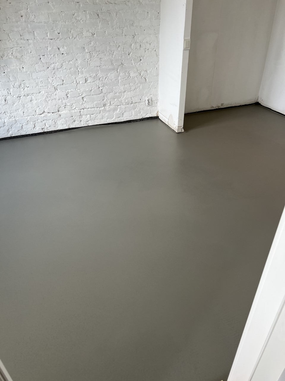 Świeżo wylana, szara posadzka w pomieszczeniu z białą, ceglaną ścianą i widocznym gniazdkiem elektrycznym.