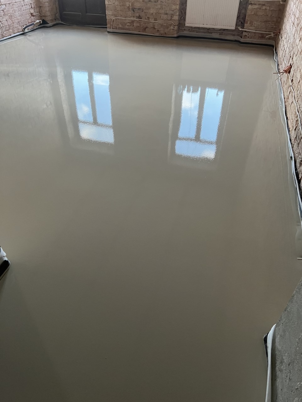 Świeżo wylana, gładka, jasnoszara posadzka z widocznymi odbiciami okien w jej lustrzanej powierzchni, ściany z surowej cegły w tle.