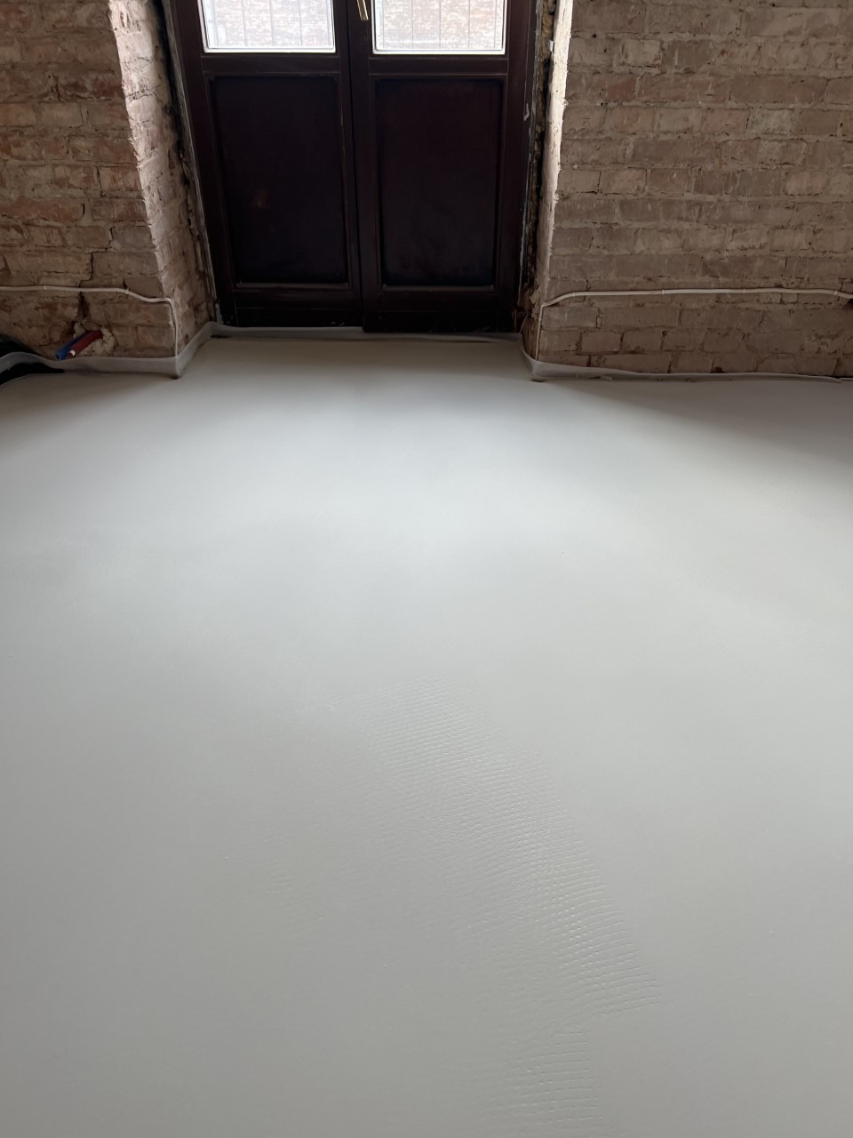 Świeżo wylana, gładka, jasnoszara podłoga w pomieszczeniu z ceglanymi ścianami i ciemnymi drzwiami balkonowymi. Widoczne ślady po wałku na powierzchni.