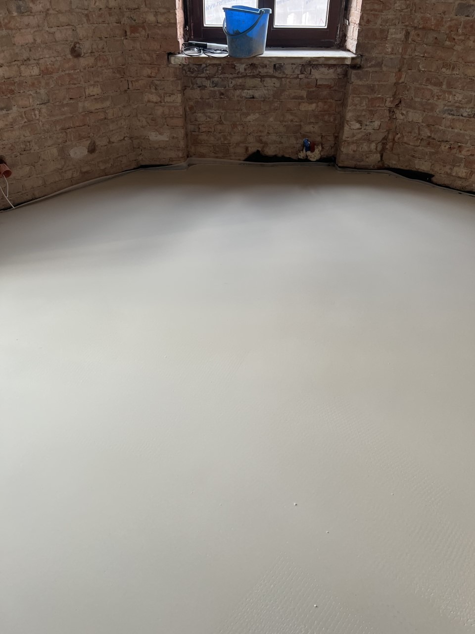 Świeżo wylana, jasna posadzka w pomieszczeniu z ceglanymi ścianami i oknem, widoczne ślady po narzędziach i wiaderko na parapecie.