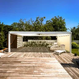 Nowoczesna, biała konstrukcja z drewnianymi elementami na drewnianym tarasie, rzucająca cienie, z widocznym zielonym otoczeniem i błękitnym niebem.