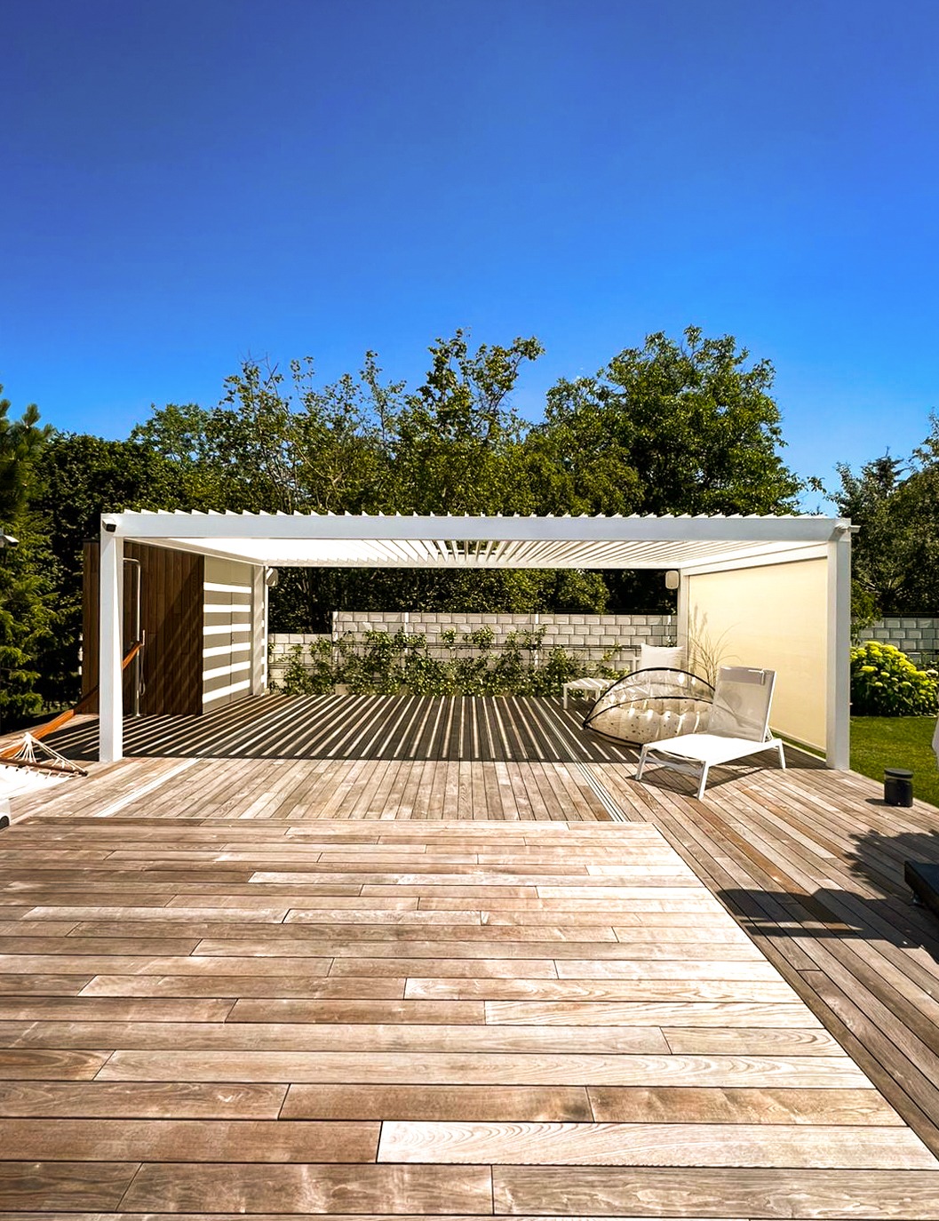 Nowoczesna, biała konstrukcja z drewnianymi elementami na drewnianym tarasie, rzucająca cienie, z widocznym zielonym otoczeniem i błękitnym niebem.