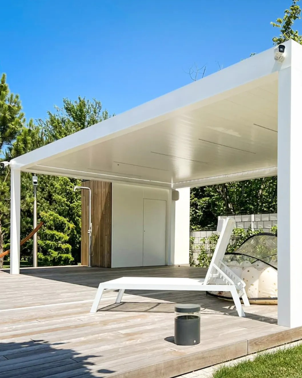 Nowoczesna, biała pergola z płaskim dachem na drewnianym tarasie z leżakiem, prysznicem zewnętrznym i kamerą monitoringu.