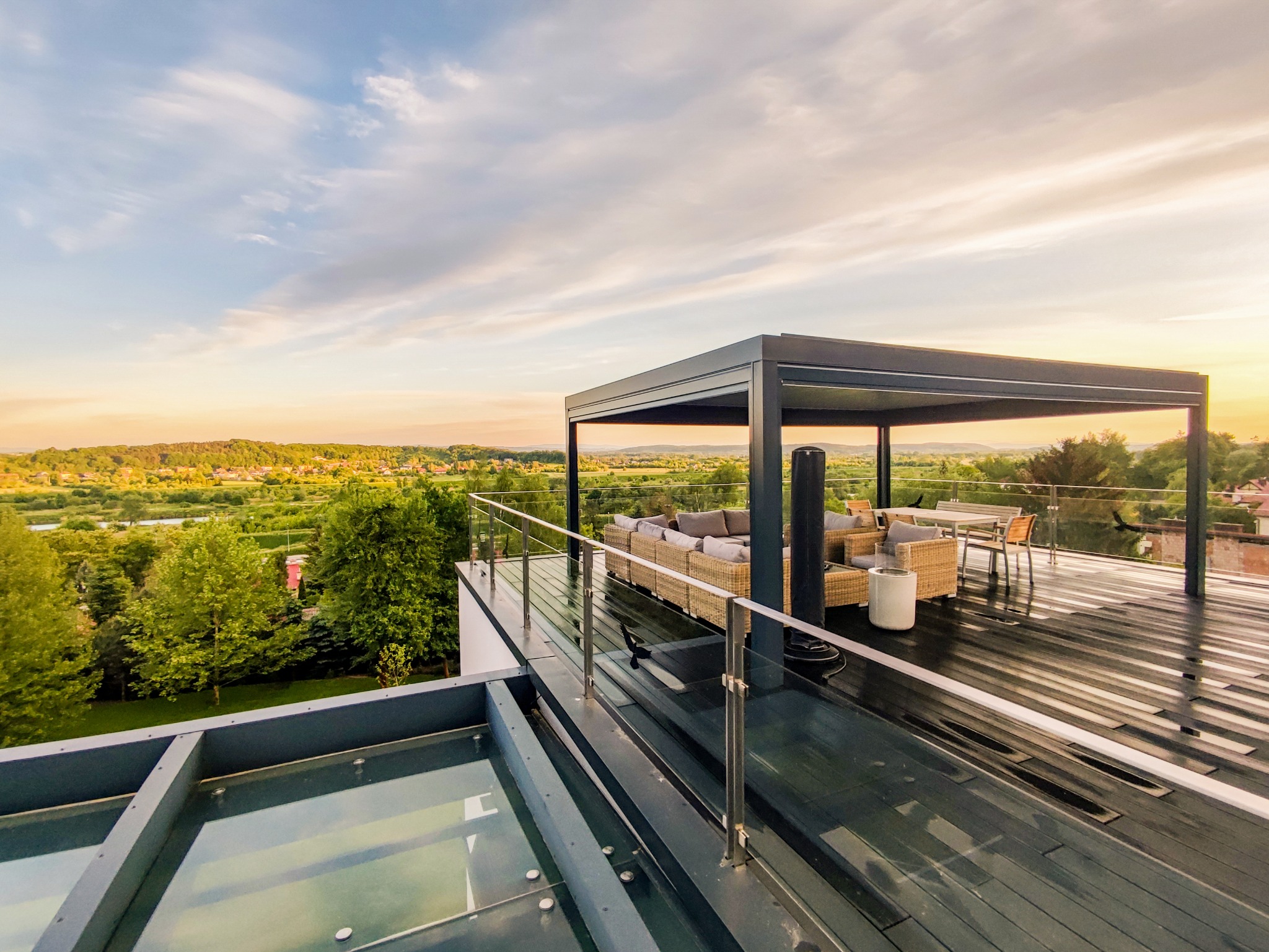 Nowoczesna altana z metalową konstrukcją na tarasie z widokiem na zielony krajobraz w promieniach zachodzącego słońca, wyposażona w rattanowe meble wypoczynkowe i szklaną balustradę.