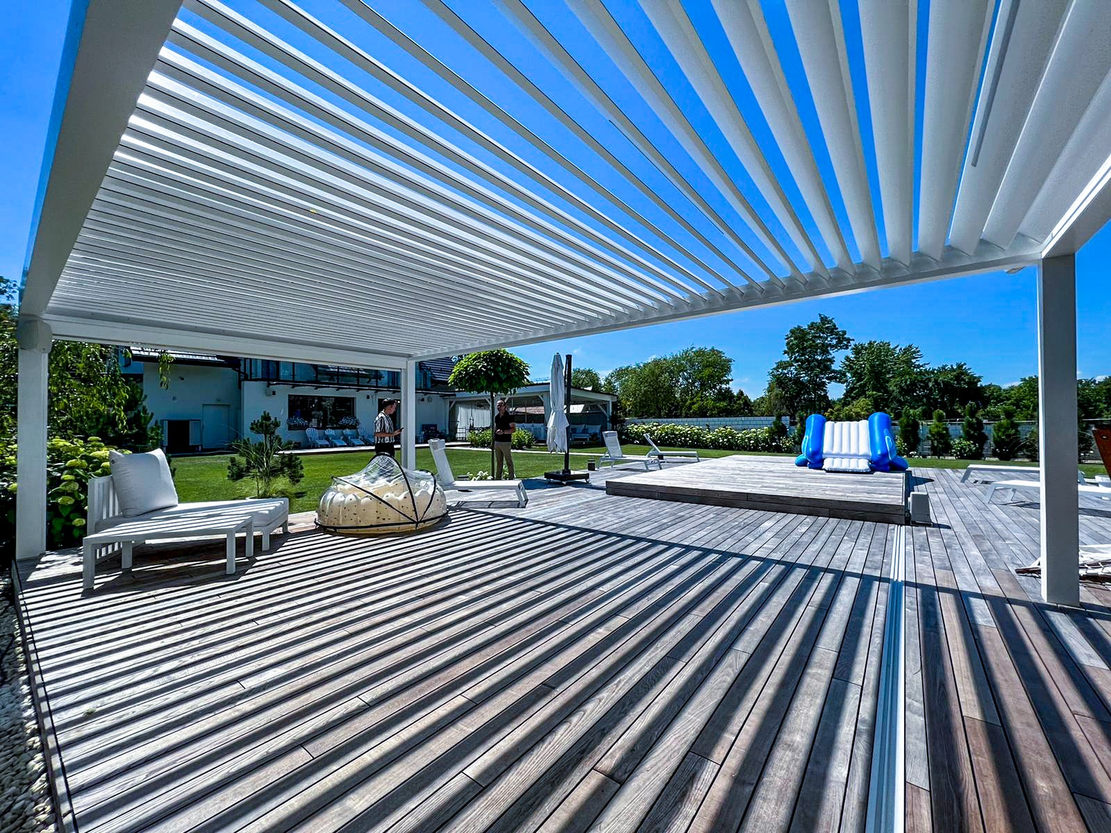 Nowoczesna, biała pergola z regulowanymi lamelami na drewnianym tarasie z widokiem na ogród, basen i dom.