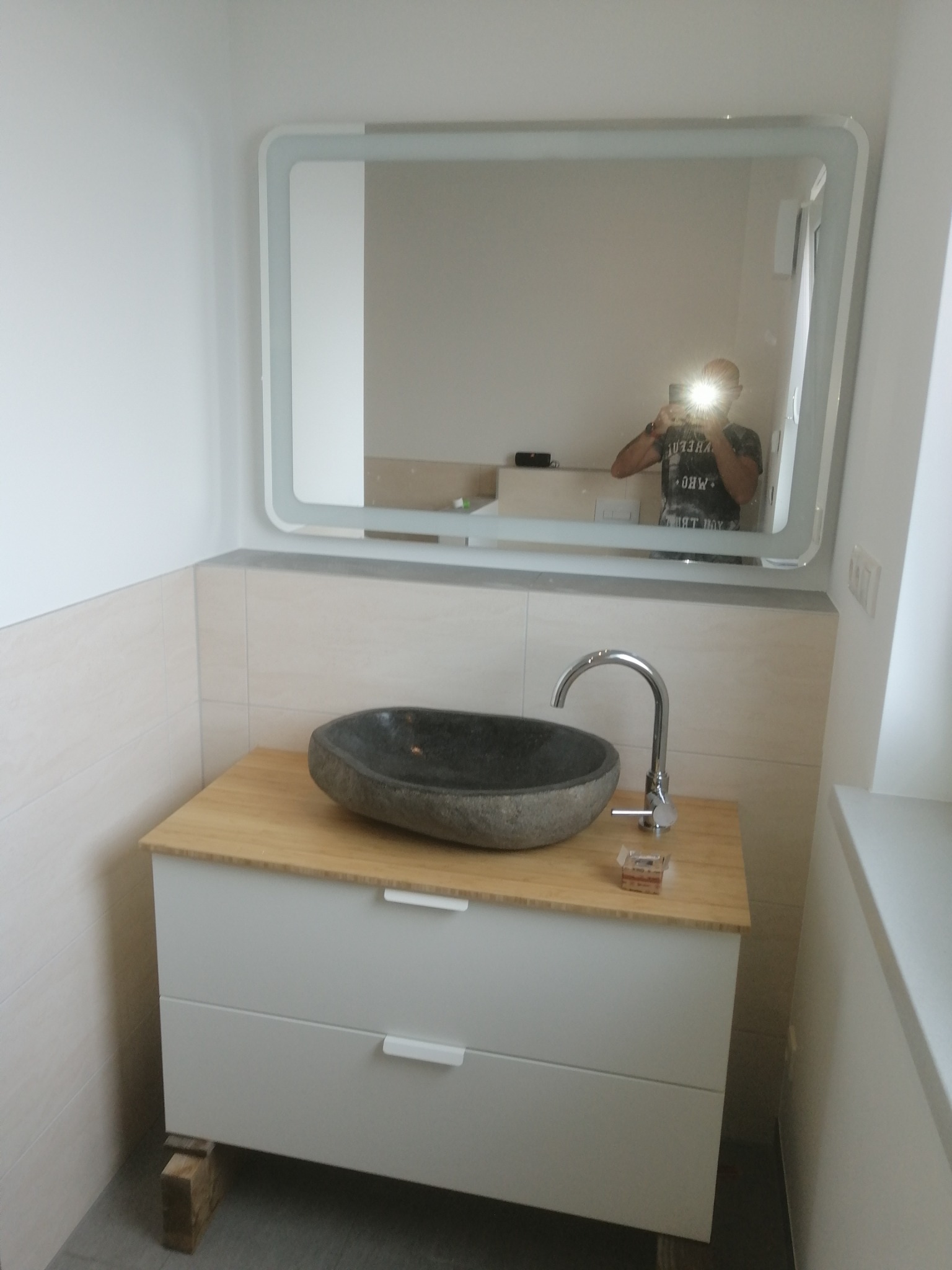 Umywalka z kamienia na szafce z drewnianym blatem w wykończonej łazience, widoczne lustro z podświetleniem i odbicie fotografa.