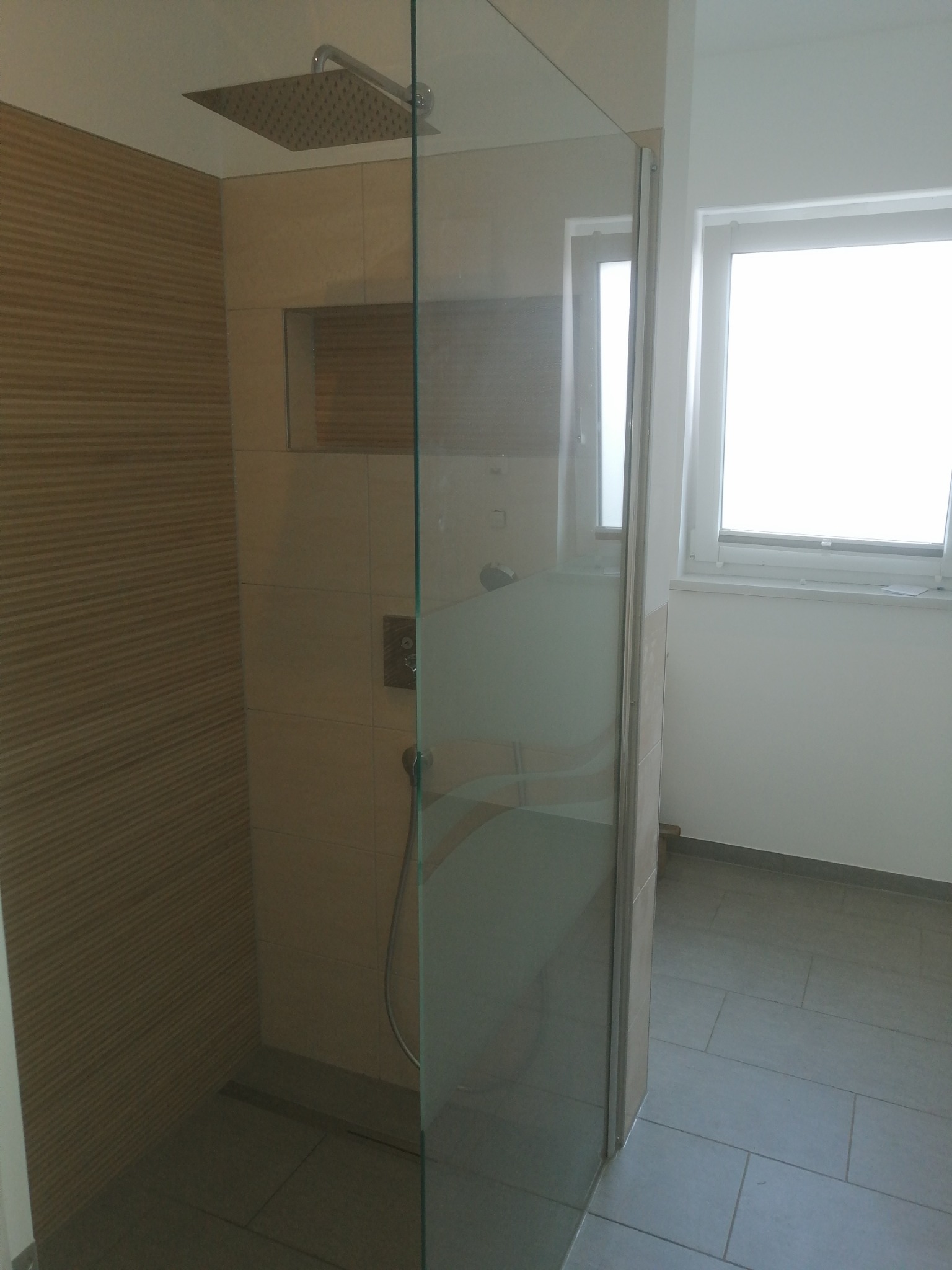 Nowoczesna kabina prysznicowa z deszczownicą i szklaną ścianką, wykończona beżowymi płytkami z poziomym wzorem, widok na okno z białymi ramami.