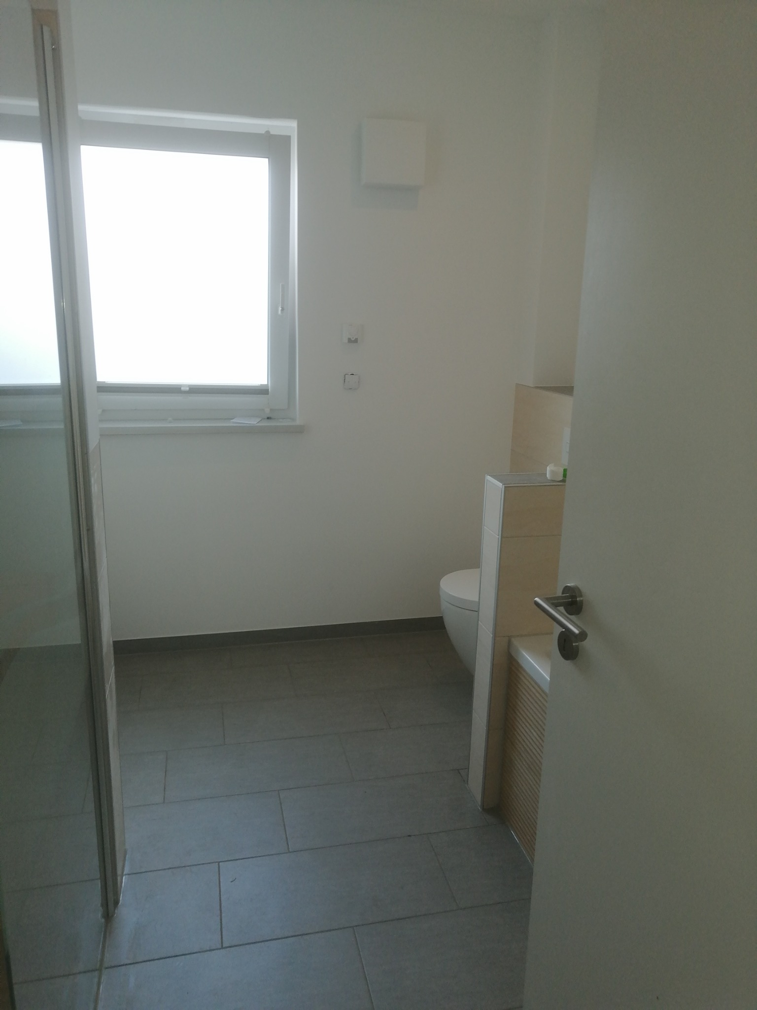 Minimalistyczna, jasna łazienka w trakcie wykańczania, widoczna toaleta, szare płytki podłogowe i fragment otwartych drzwi z klamką.