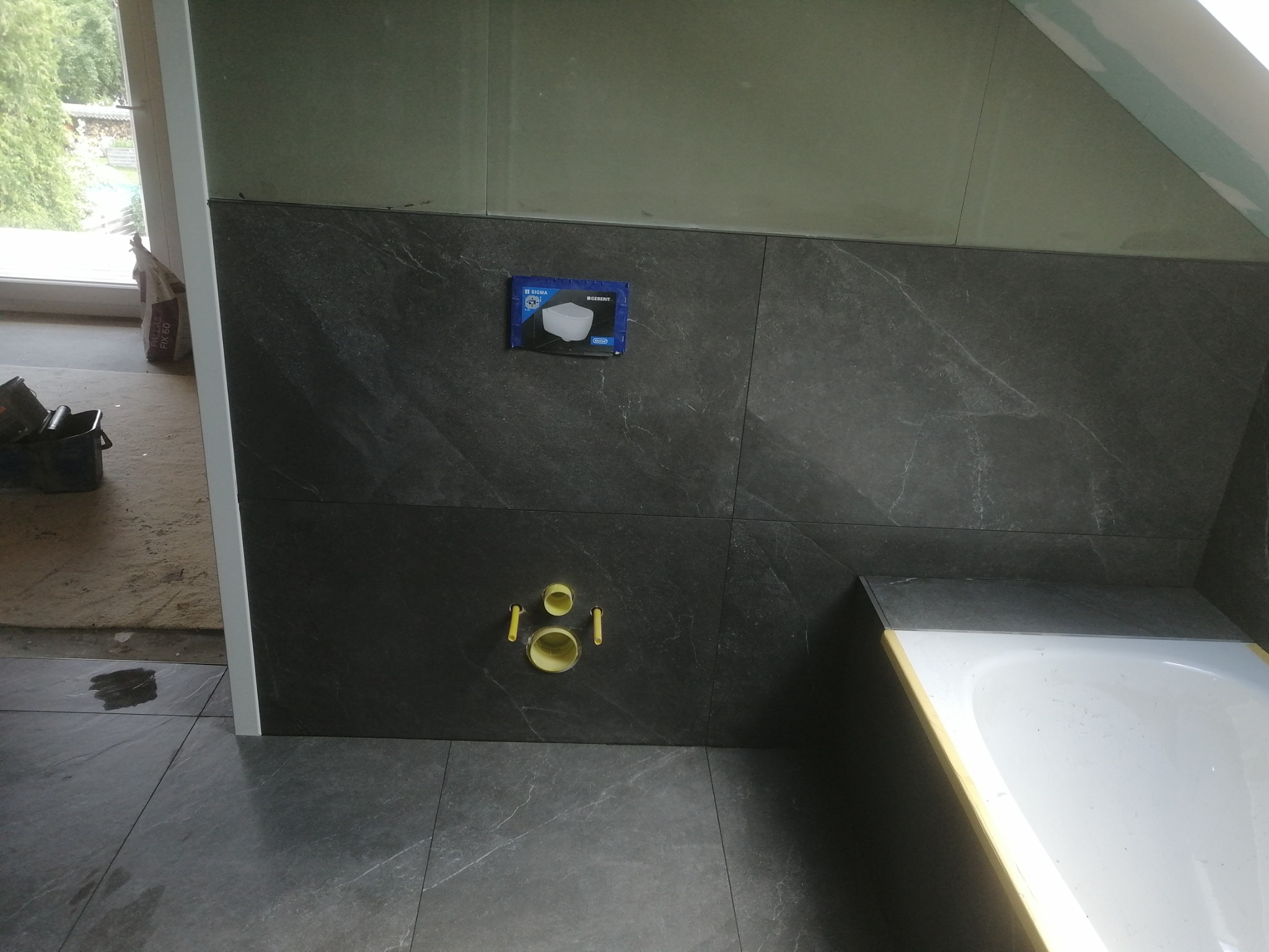 Częściowo wykończona łazienka w poddaszu z szarymi płytkami imitującymi marmur na ścianach i podłodze, widoczne żółte rury odpływowe, fragment wanny i opakowanie po stelażu podtynkowym.