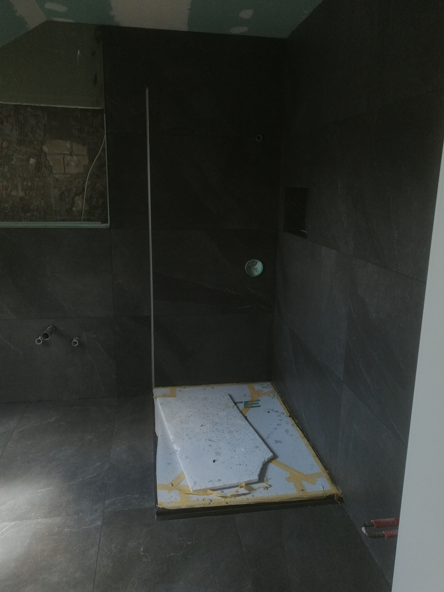 Ciemnoszara łazienka w trakcie remontu z widocznymi płytkami, przygotowanym brodzikiem zabezpieczonym kartonem i taśmą oraz fragmentem instalacji wodnej.
