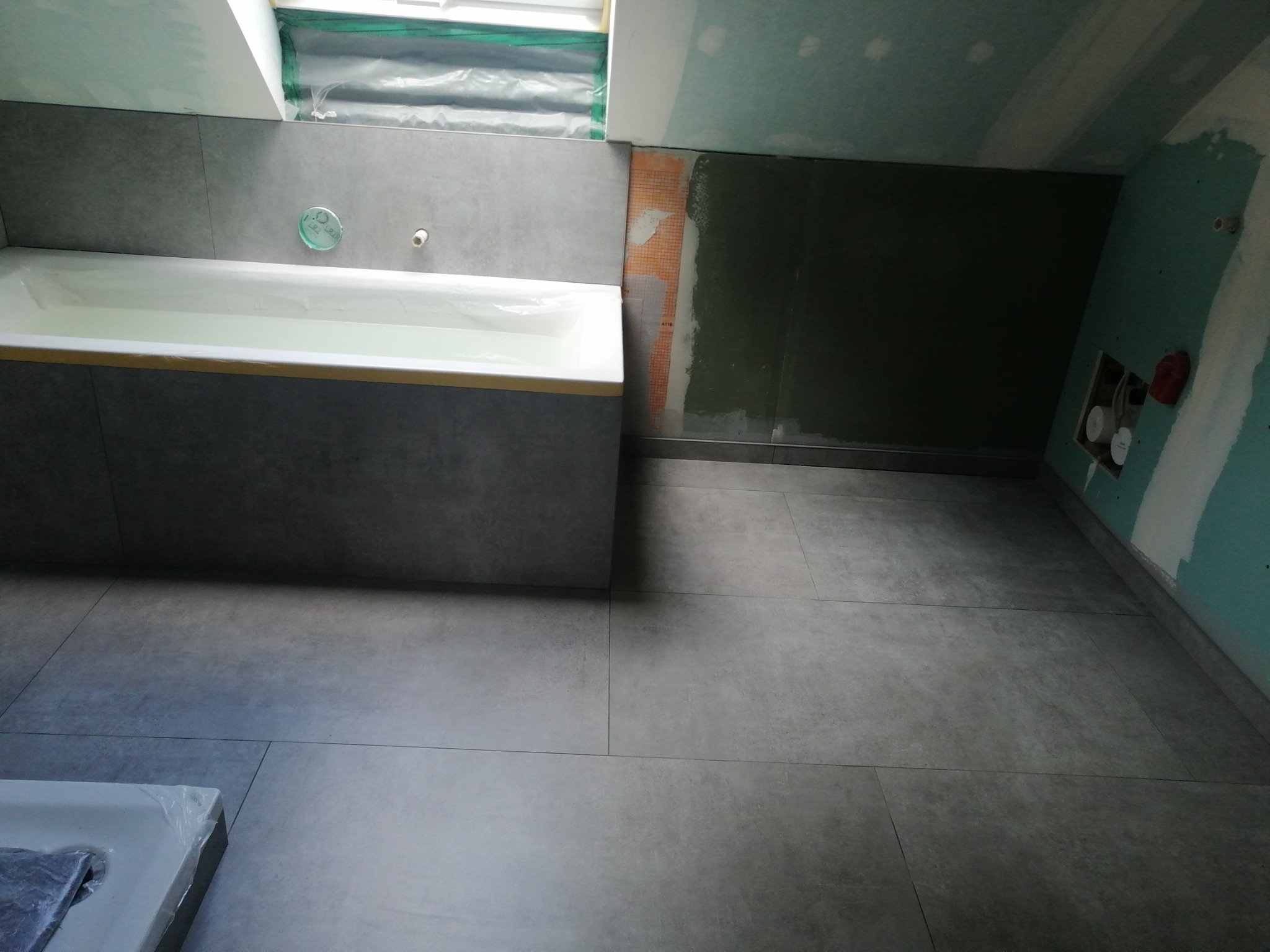 Łazienka w trakcie remontu: szare płytki podłogowe i ścienne, biała wanna częściowo zabudowana, ściany częściowo przygotowane do dalszych prac wykończeniowych, widoczne instalacje.