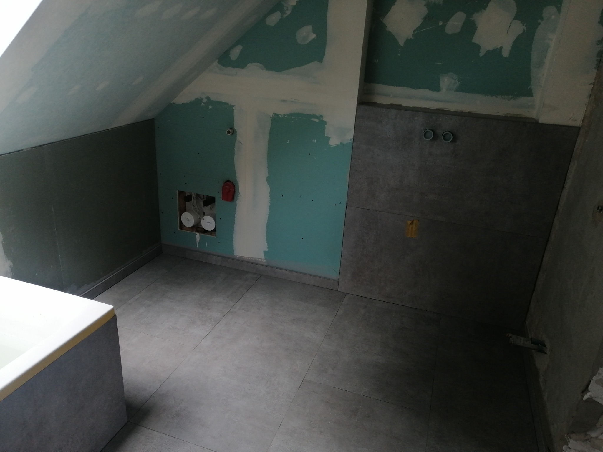 Łazienka w trakcie remontu: szare płytki na podłodze i ścianach, fragment wanny, ściany częściowo pokryte płytami gipsowo-kartonowymi z widocznymi śladami szpachlowania i instalacją hydrauliczną.