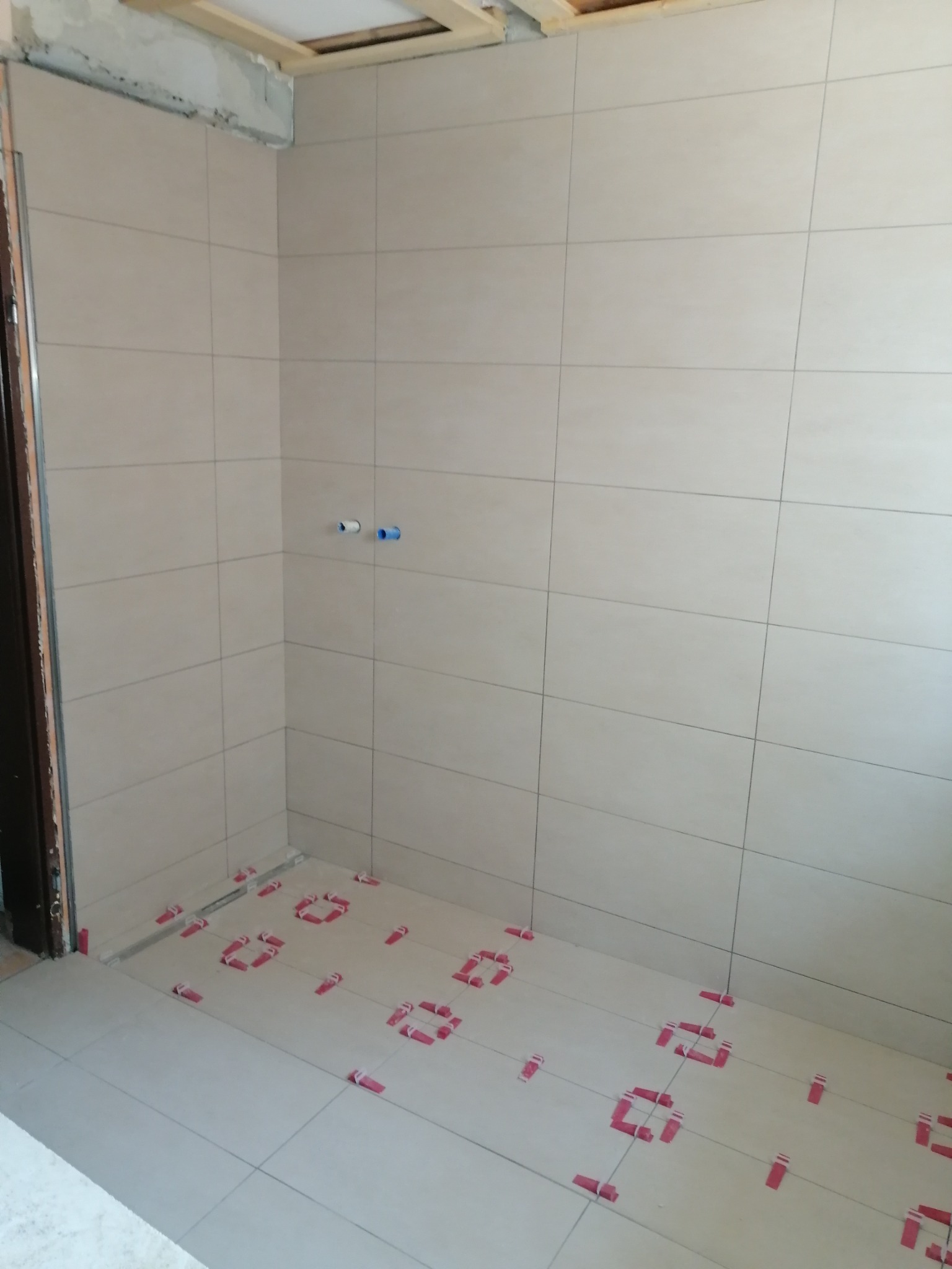 Świeżo ułożone, beżowe płytki podłogowe i ścienne w łazience, z czerwonymi klipsami poziomującymi, widoczne rury instalacji wodnej i surowy stan wykończenia sufitu.