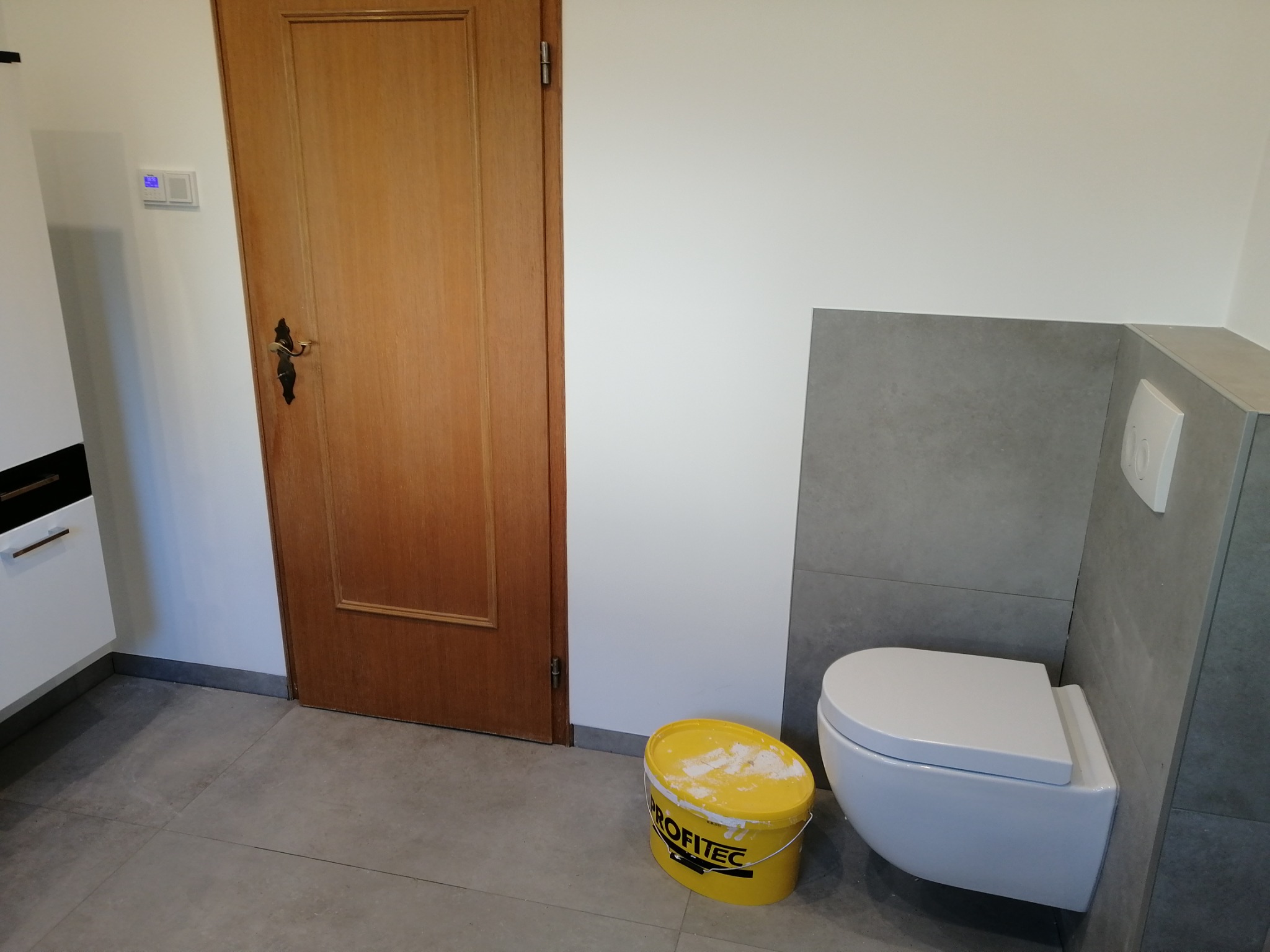 Świeżo pomalowana łazienka z wiszącą toaletą, szarymi płytkami i żółtym wiadrem po farbie marki Profitec.