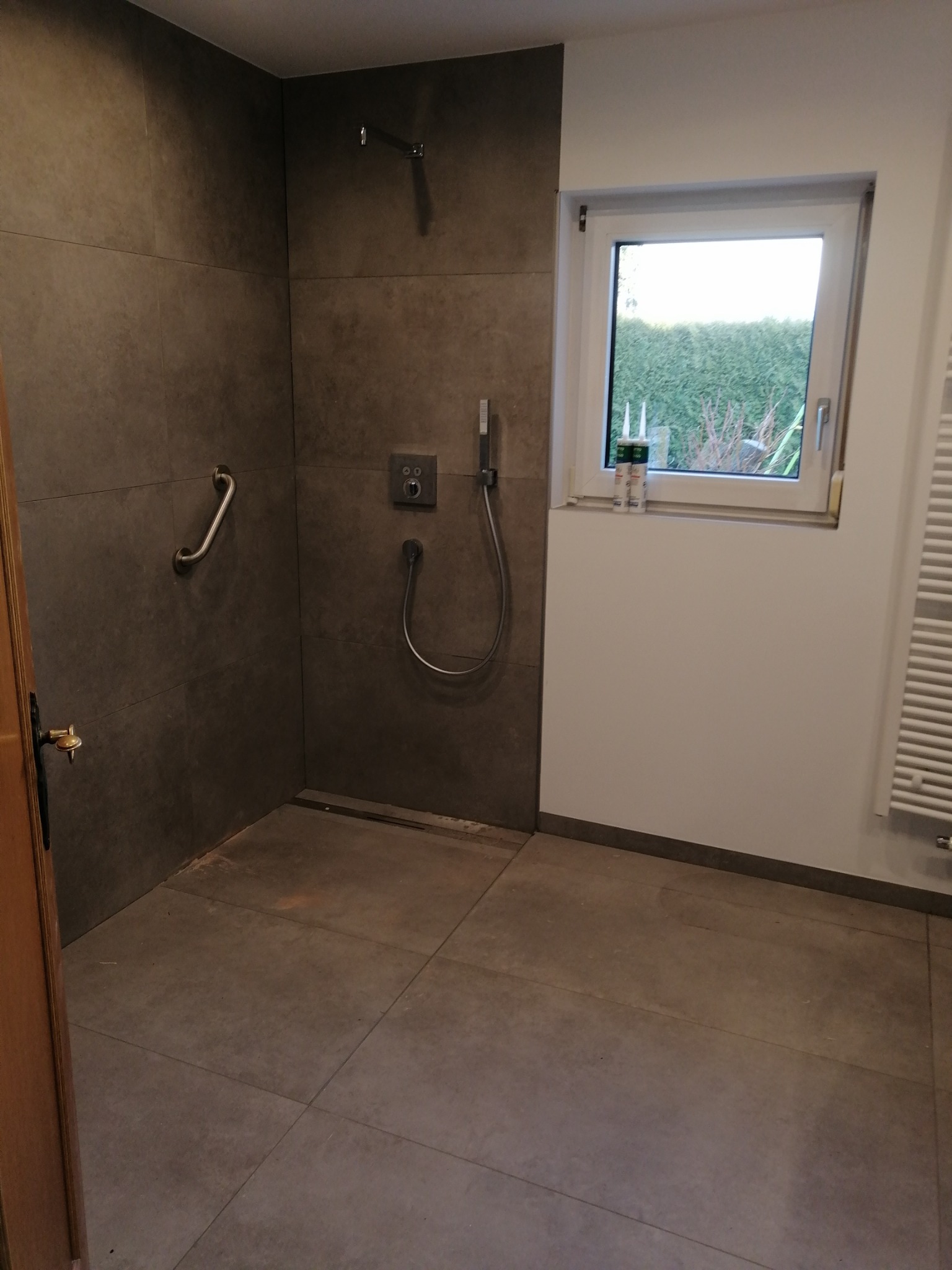 Nowoczesna kabina prysznicowa z szarymi płytkami, uchwytem dla niepełnosprawnych i odpływem liniowym, widok na okno z uszczelniaczami na parapecie.