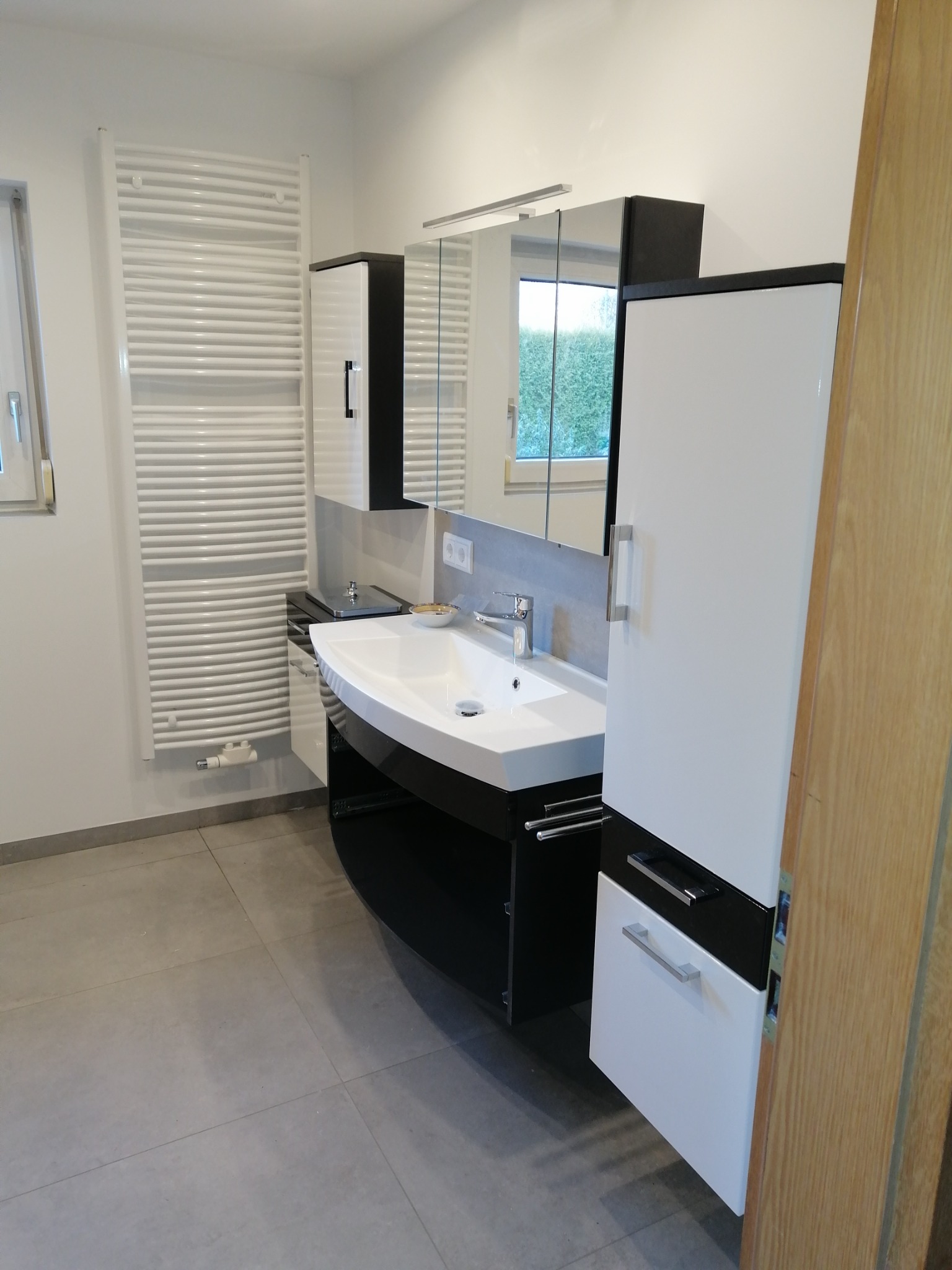 Nowoczesna łazienka z umywalką nablatową, szafką w kolorze czarno-białym, lustrem z oświetleniem, grzejnikiem drabinkowym i szarą podłogą z płytek.