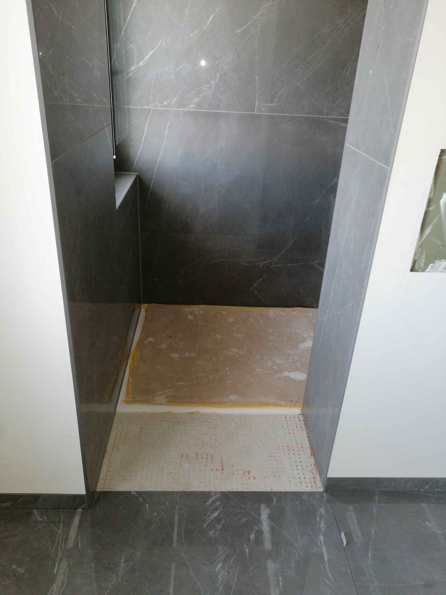 Szara łazienka z marmurowymi płytkami w trakcie remontu, widoczne przygotowane podłoże pod prysznic i częściowo ułożona podłoga.
