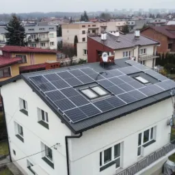 Biały dom jednorodzinny z ciemną dachówką i panelami słonecznymi, widoczne okna dachowe oraz drabina oparta o ścianę, w tle zabudowa miejska.