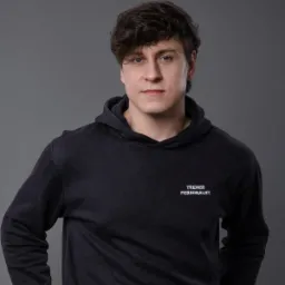 Portret mężczyzny w ciemnej bluzie z kapturem, na której widnieje napis 'TRENER PERSONALNY', na szarym tle.