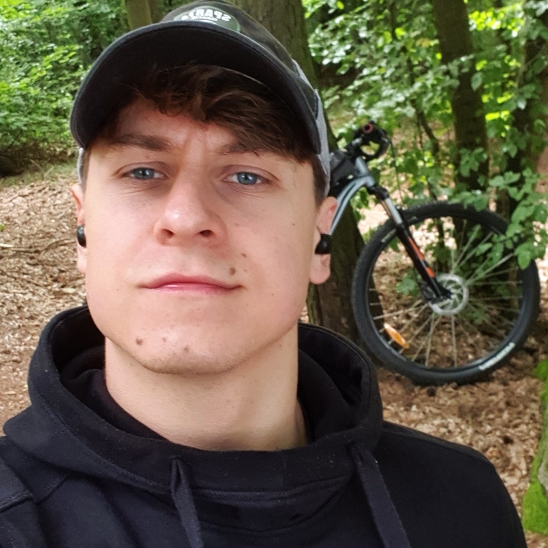 Młody mężczyzna w czarnej bluzie z kapturem i czapce z daszkiem, robiący selfie na tle roweru górskiego w lesie.