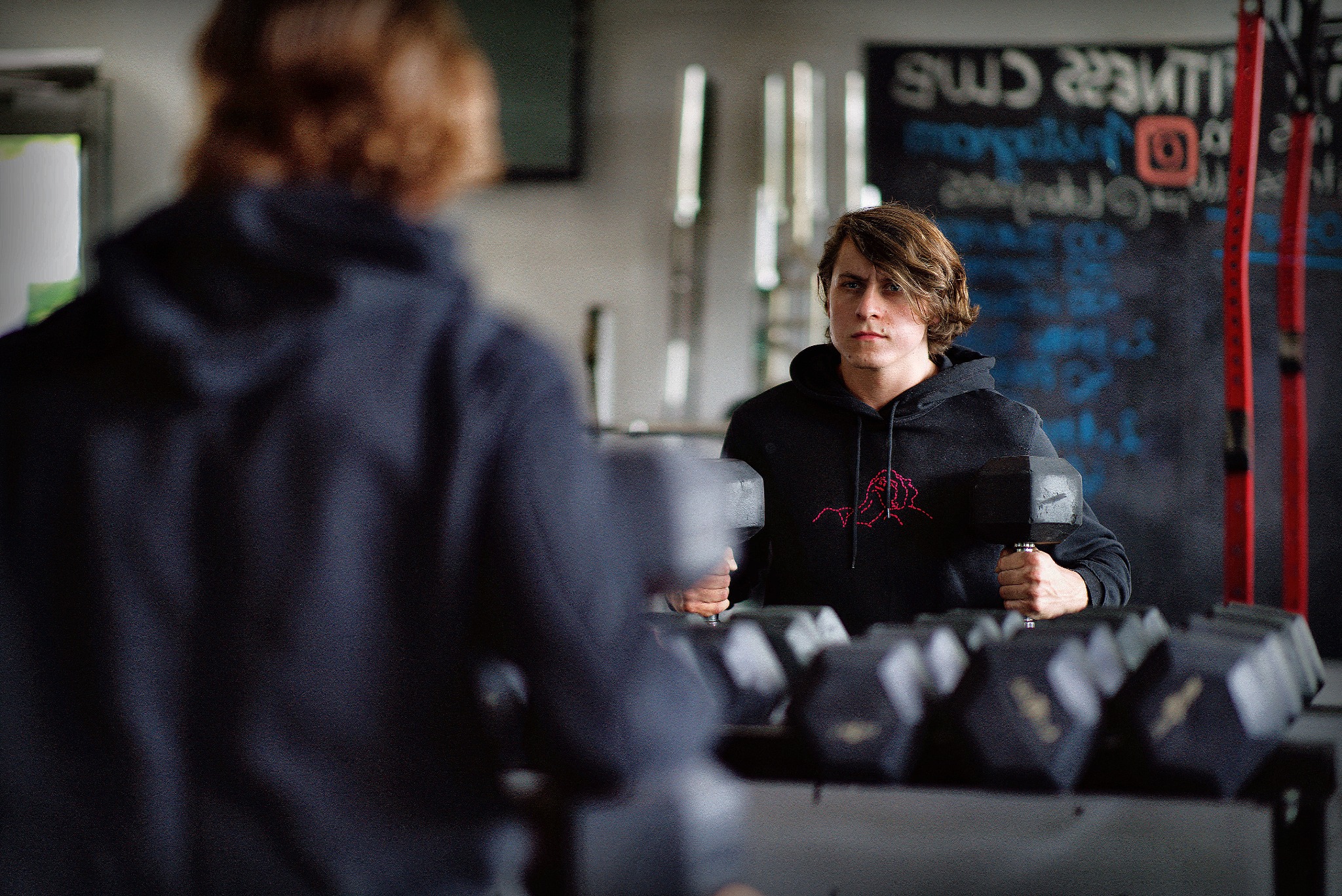 Młody mężczyzna w czarnej bluzie z kapturem, z czerwonym wzorem, trzyma ciężarek w siłowni, widoczny w lustrze z rzędem ciężarków.