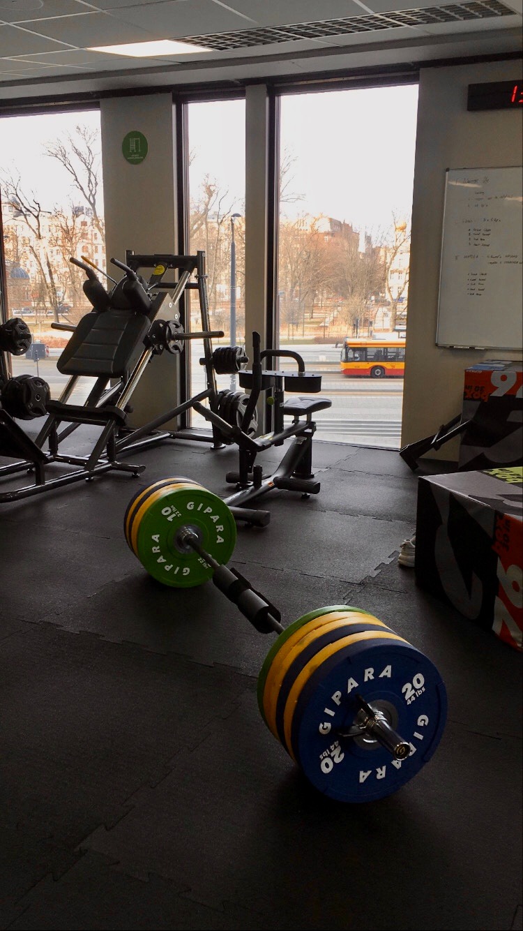 Ciężka sztanga z obciążeniami GIPARA na podłodze w siłowni, w tle maszyny do ćwiczeń i widok na miasto z autobusem komunikacji miejskiej.