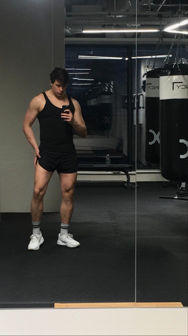 Mężczyzna w czarnym tank topie i spodenkach pozuje w lustrze siłowni, widoczne worki treningowe w tle.