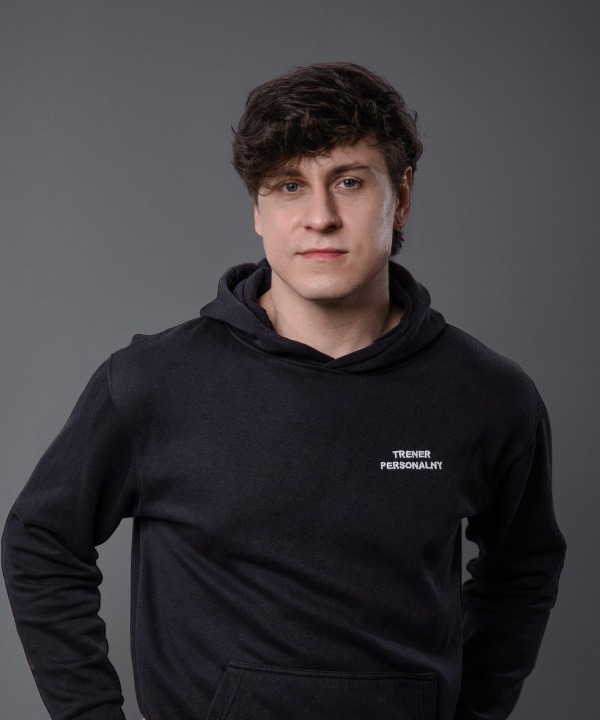 Portret mężczyzny w ciemnej bluzie z kapturem, na której widnieje napis 'TRENER PERSONALNY', na szarym tle.