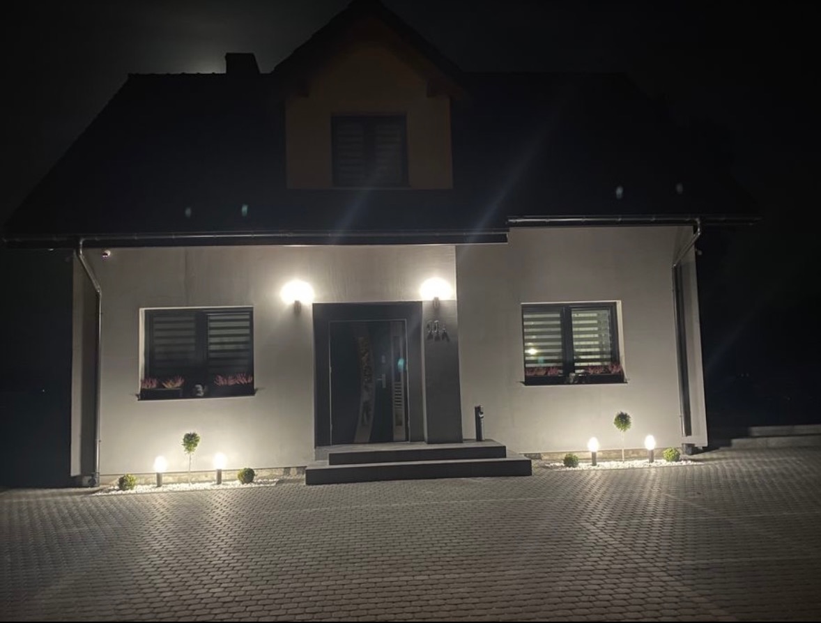 Nowoczesny dom jednorodzinny nocą, oświetlony lampami wzdłuż ścieżki z kostki brukowej i nad wejściem, z widocznymi donicami z kwiatami w oknach.