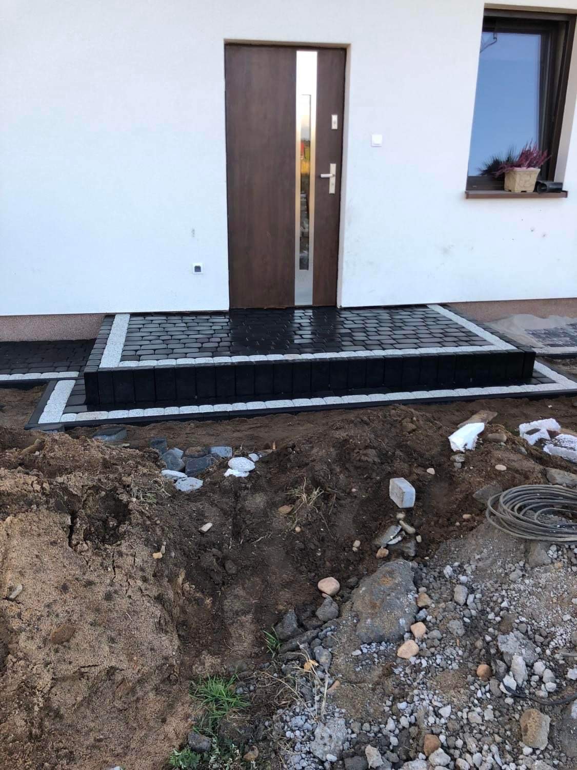 Ułożony podest z czarnej kostki brukowej z białymi obrzeżami przed wejściem do domu, widoczny fragment niedokończonego terenu z ziemią i kamieniami.