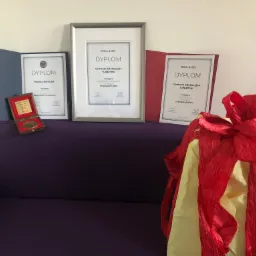 Dyplomy i nagrody na fioletowej sofie, w tym dyplomy dla Centrum Edukacyjnego Tulipanów i Moniki Bronickiej, z dekoracyjnym czerwono-żółtym prezentem z bibuły z przodu.