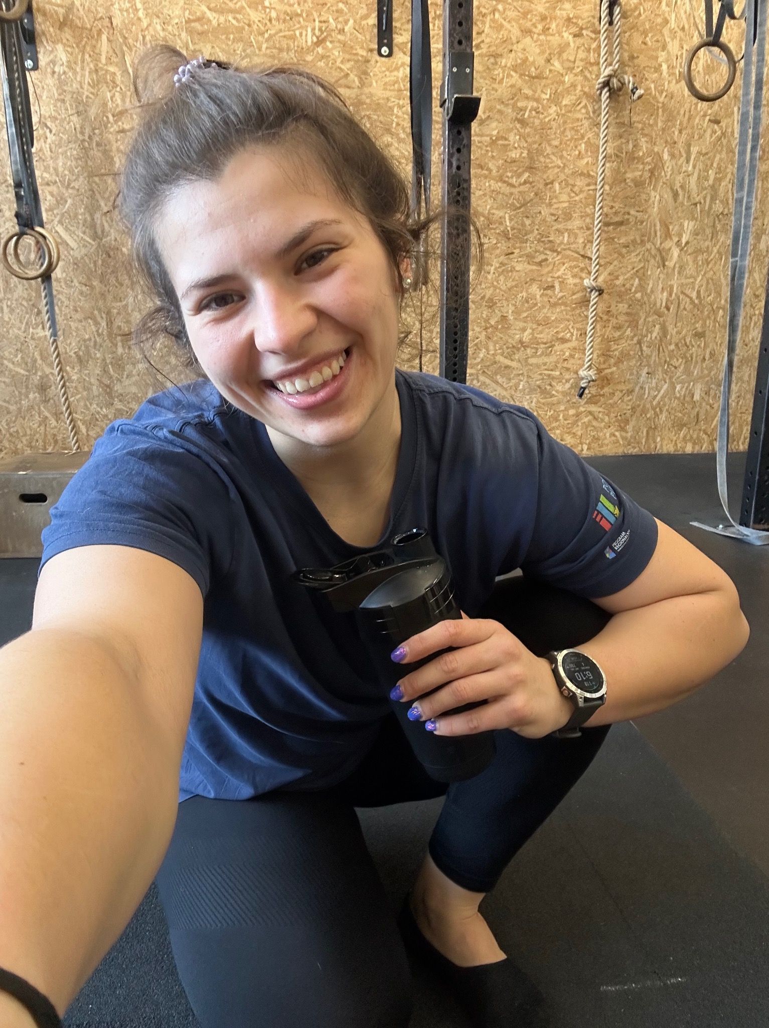 Uśmiechnięta kobieta w sportowym stroju, w siłowni, trzymająca bidon, z widocznym zegarkiem sportowym i elementami wyposażenia siłowni w tle.